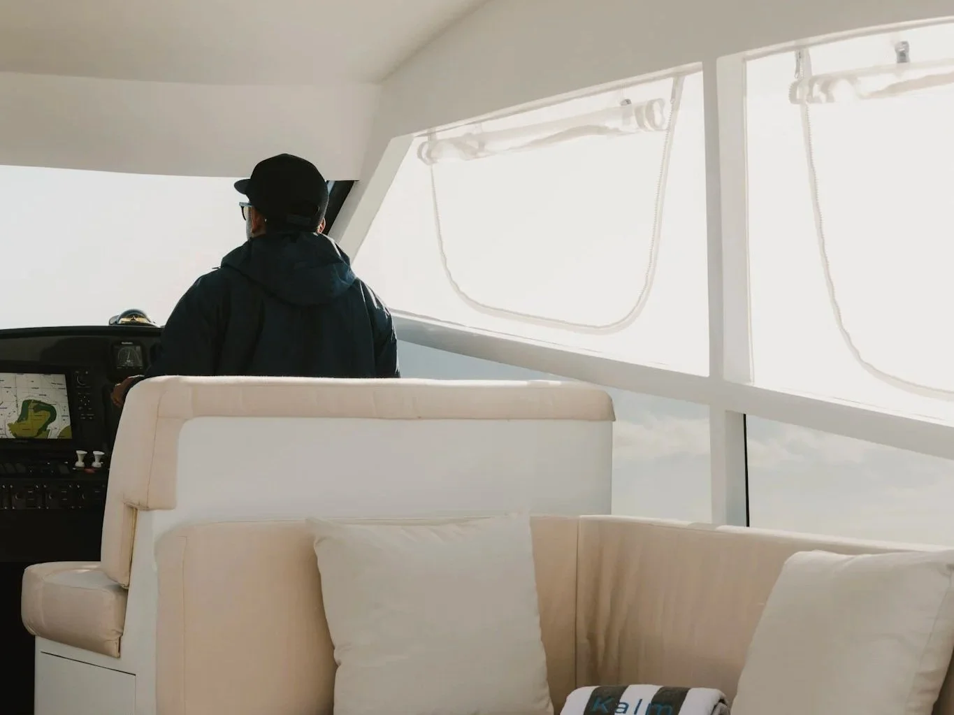 Person in a black jacket and cap inside a boat, looking out through large windows with white framing.