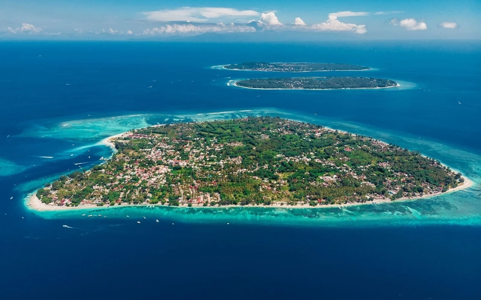 Aerial view of an island with lush green trees and residential areas, surrounded by turquoise waters and white sandy beaches.