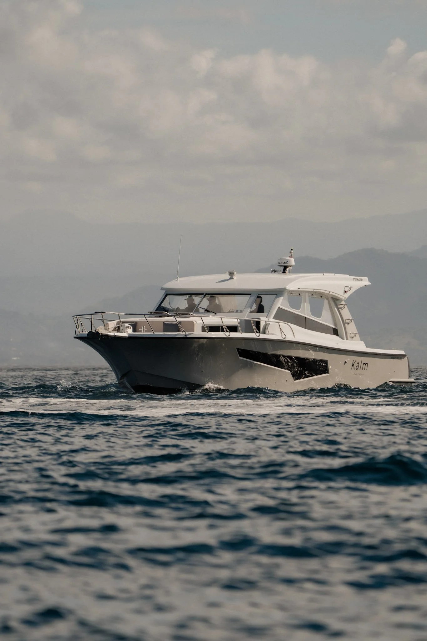 White yacht named Kalm sailing on water with mountainous landscape and cloudy sky in the background.