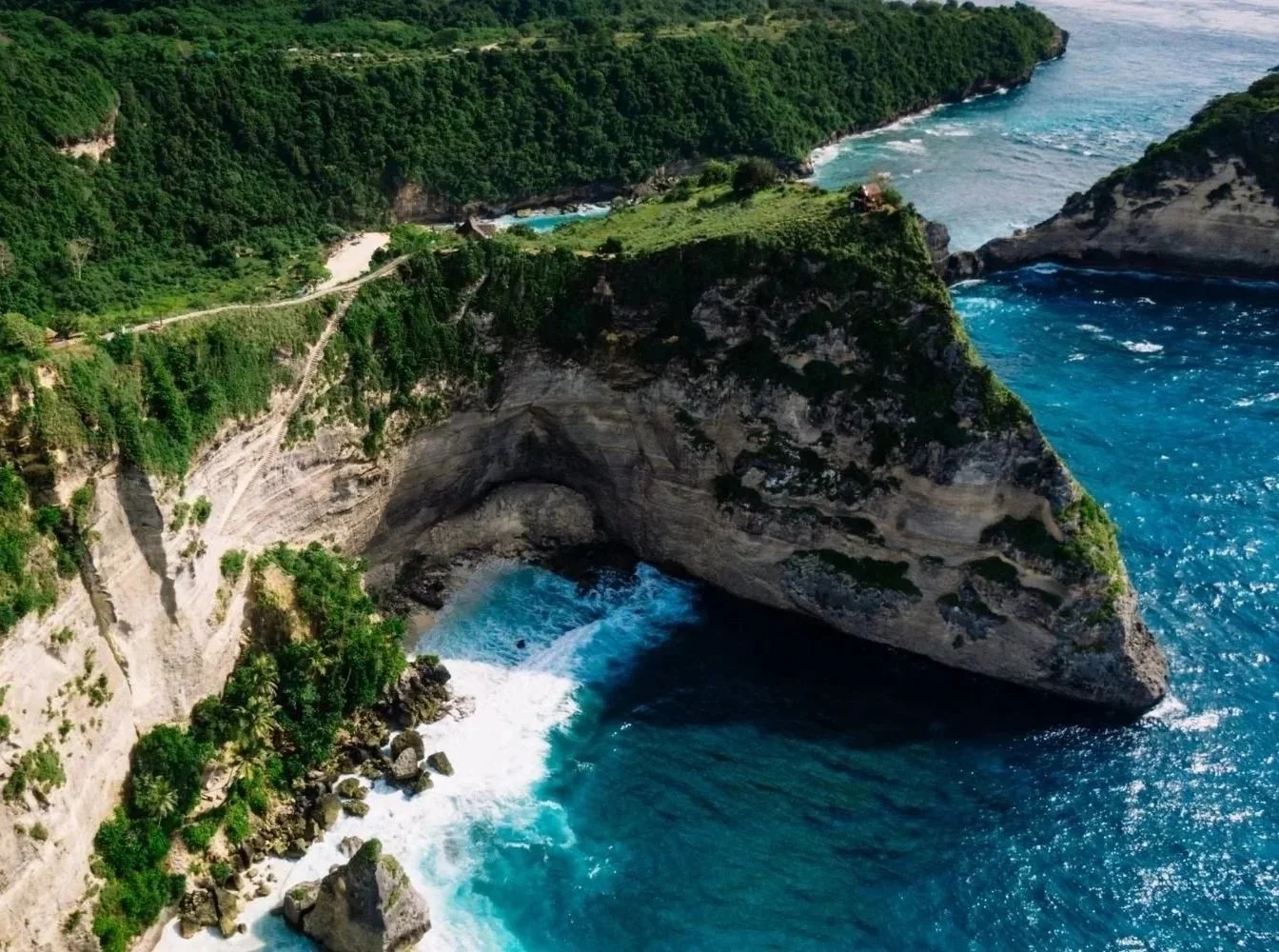 Aerial view of a natural sea arch on a rugged coastline with lush greenery, rocky cliffs, and blue ocean waves crashing underneath.