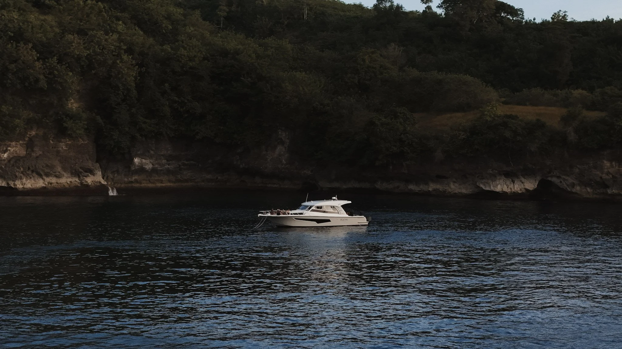 A white yacht named Kalm floating on calm dark water near a rocky shoreline with dense trees and hillside in the background during twilight or early evening.