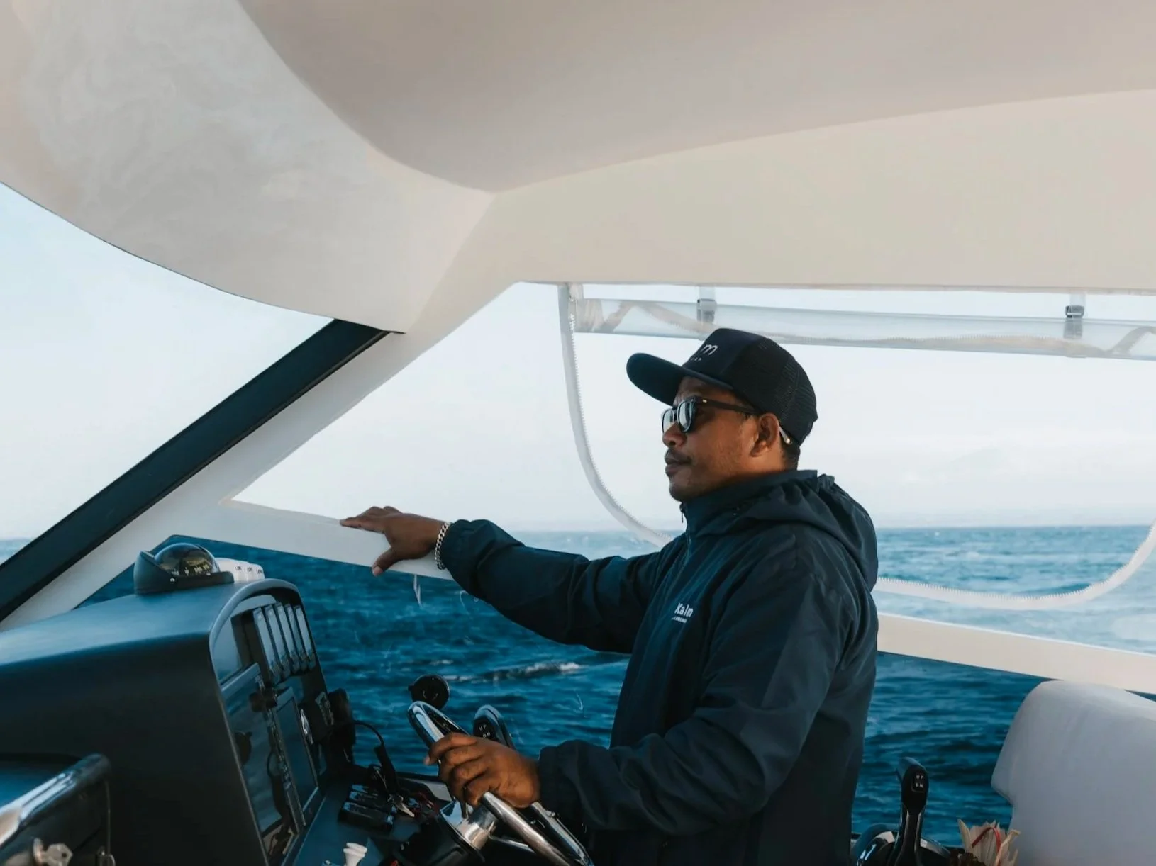 A man wearing sunglasses, a black cap, and a black jacket steering a boat on the ocean, with his right hand on the wheel and holding a smartphone in his left hand. The boat's control panel is visible in the foreground.