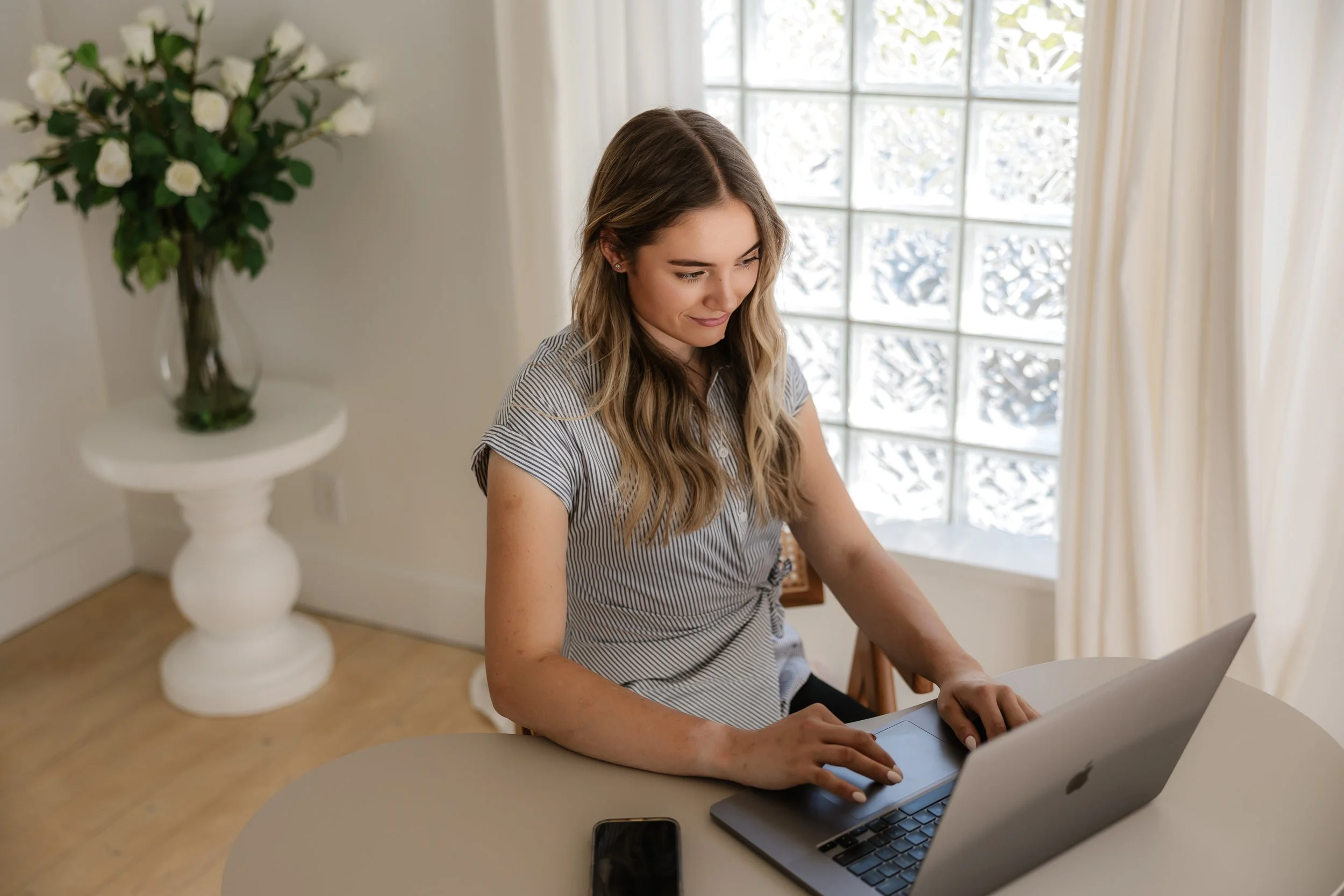 A young woman with wavy light brown hair sitting at a round table, working on a silver laptop, near a large window with white curtains, in a bright room with a decorative glass brick wall and a large vase of white roses on a white pedestal in the background.