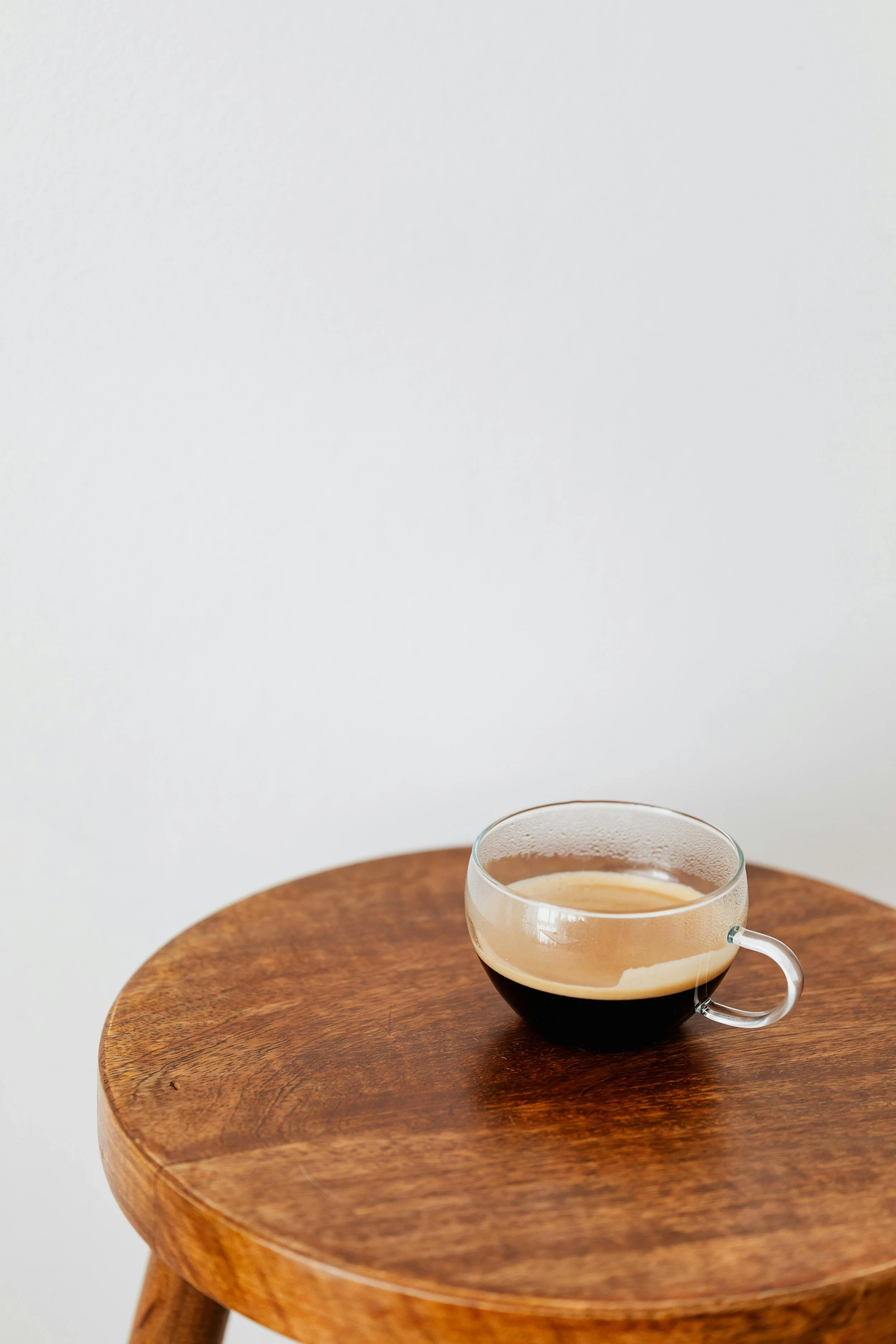 A clear glass mug filled with black coffee on a round wooden table with a white background.
