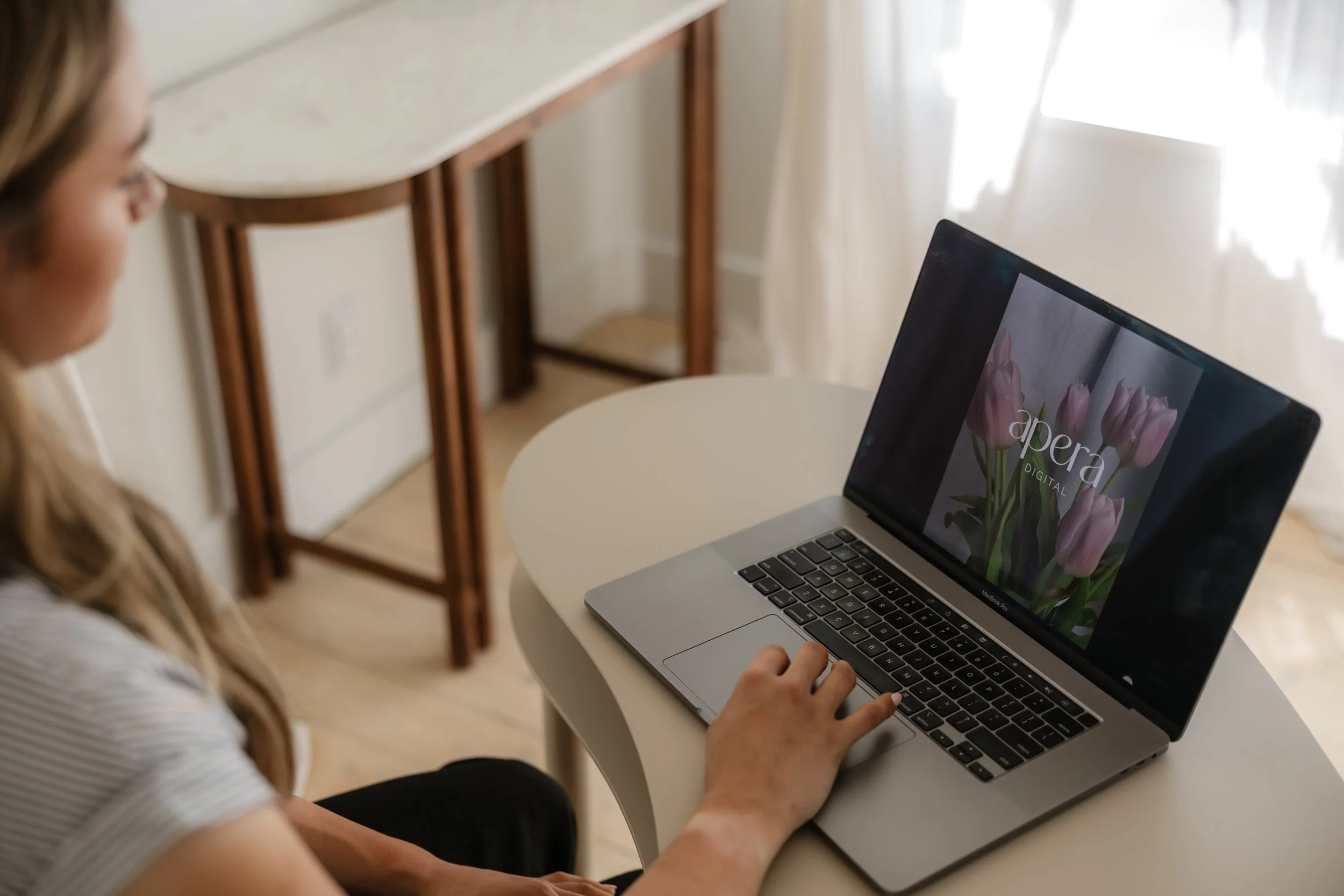A woman sitting at a white round table, looking at a MacBook laptop with a flower wallpaper on the screen, in a room with sheer white curtains, a wooden console table, and natural light.