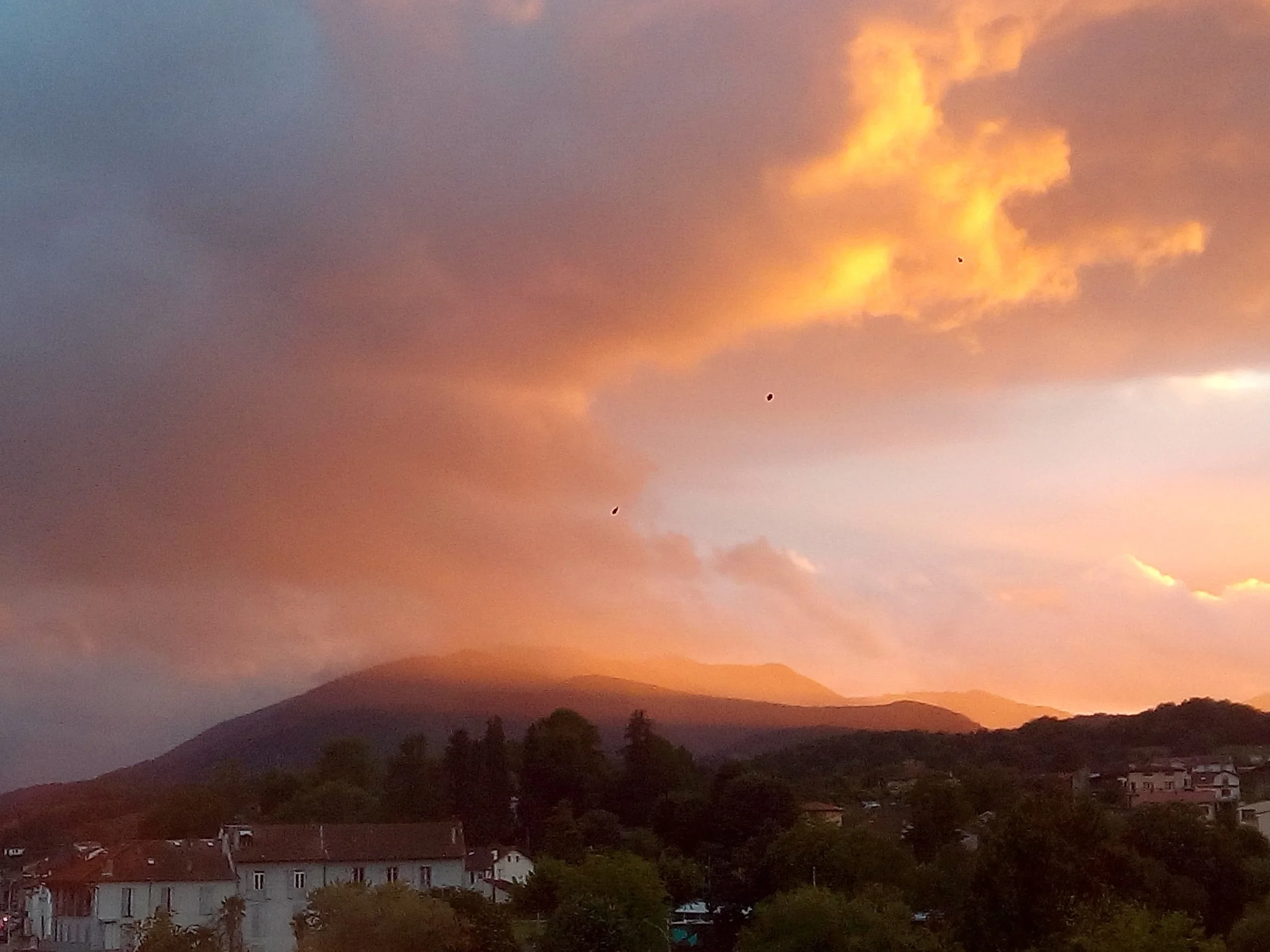 Coucher de soleil avec ciel orange et nuages, silhouettes de montagnes et villages en bas.