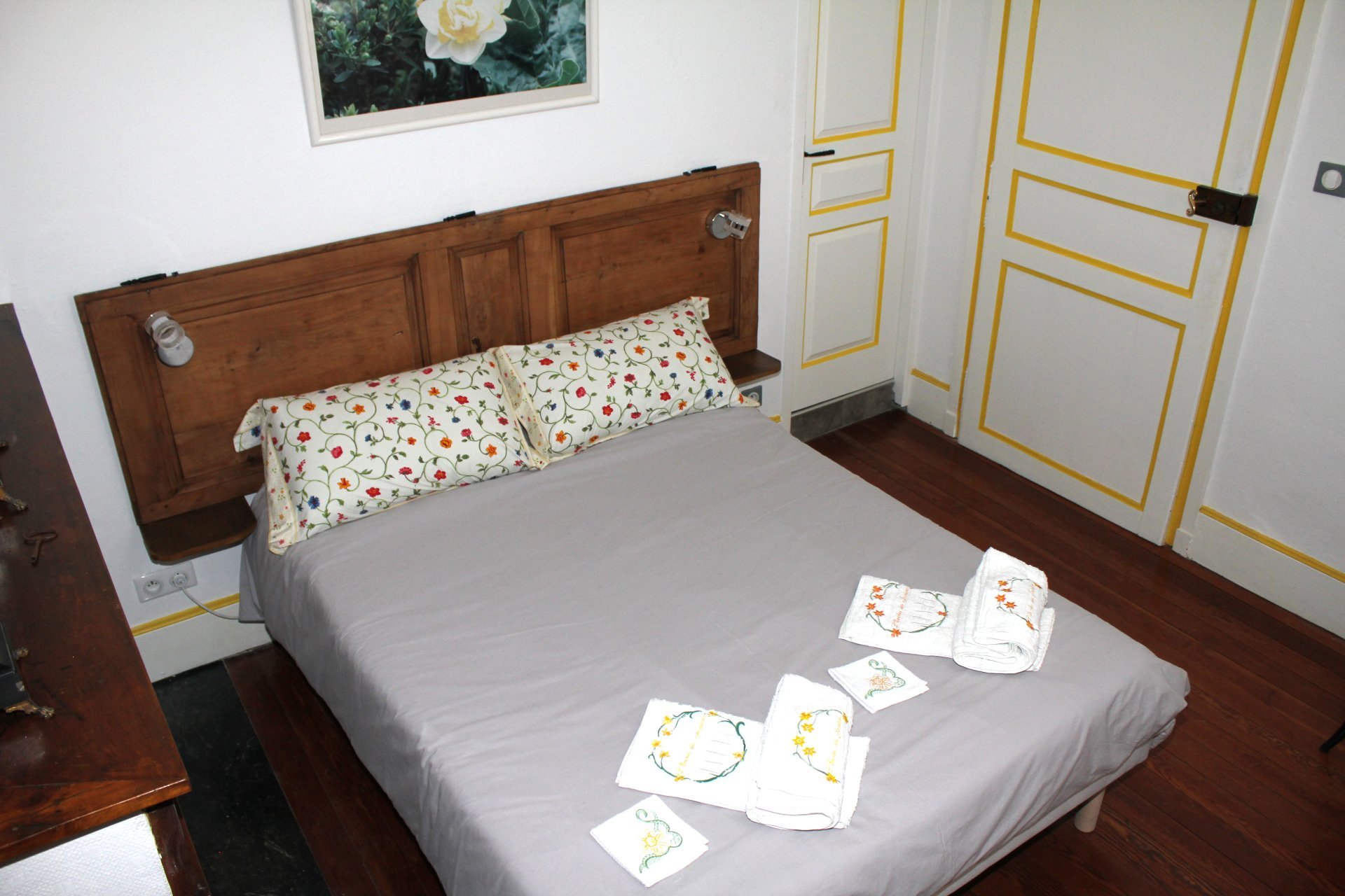 Lit avec un sommier en bois, oreillers à motifs floraux, serviettes pliées avec broderies florales posées dessus, porte blanche avec des détails jaunes dans une chambre.