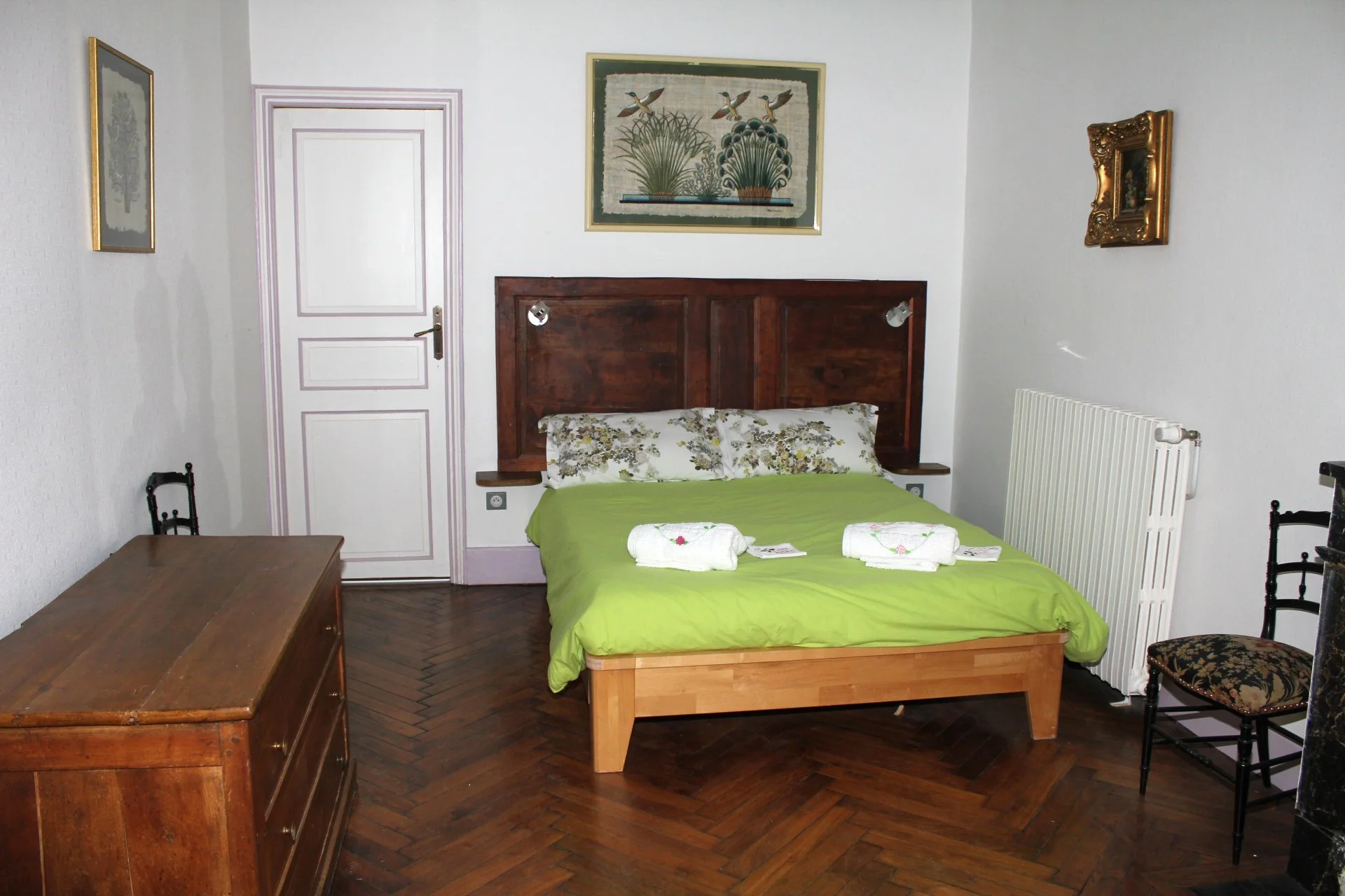 Chambre avec un lit en bois avec une tête de lit en bois foncé, draps verts, deux oreillers avec housses à motif floral, serviettes blanches pliées sur le lit, tableau de trois canards et de la végétation au-dessus du lit, porte blanche, radiateur blanc, deux lampes inclinées fixées à la tête de lit, deux chaises en bois avec rembourrage floral, table en bois.