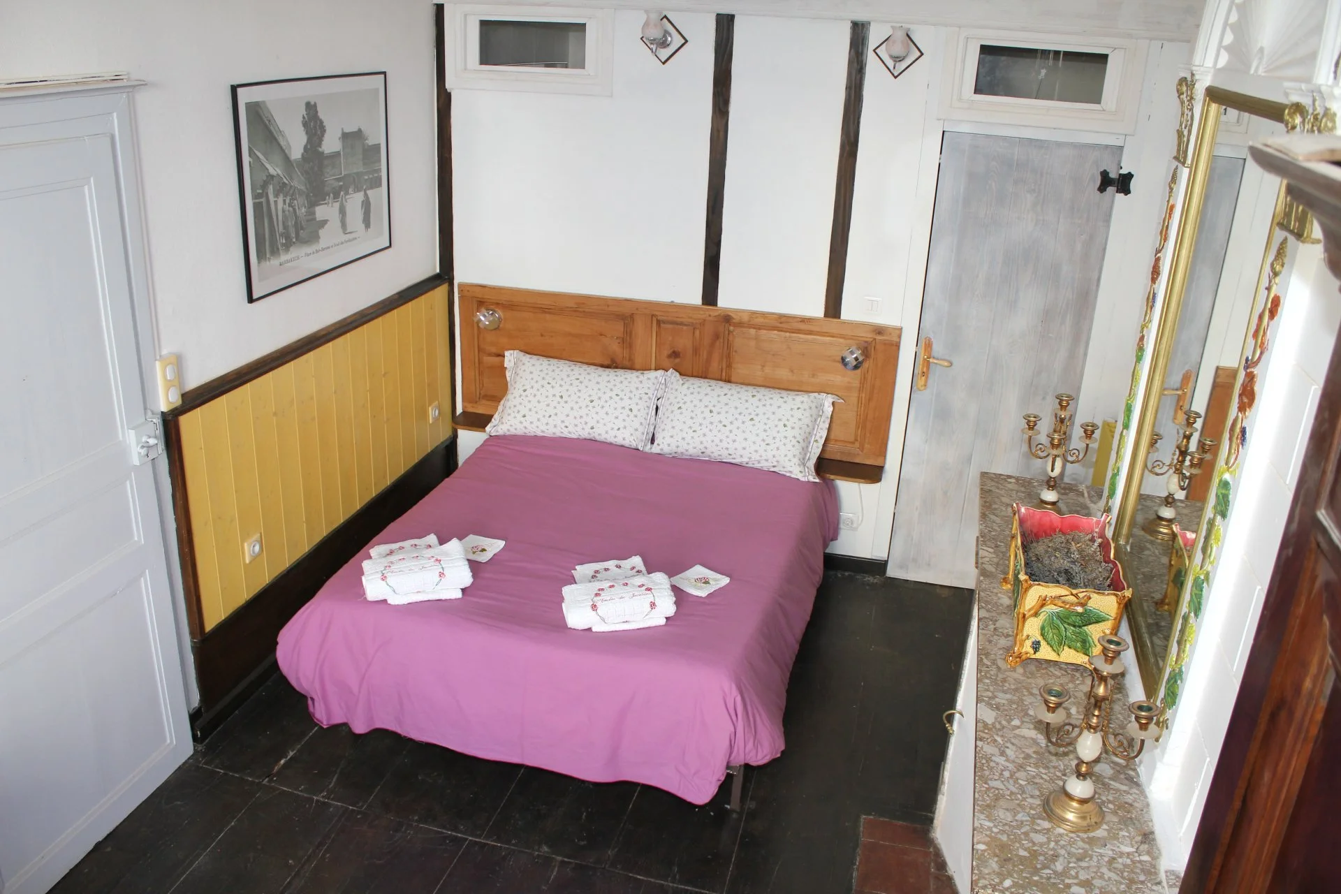 Chambre avec lit double en bois, draps roses, serviettes blanches pliées sur le lit, tableau noir et blanc sur la paroi, porte en bois gris, mur peint en blanc avec poutres verticales en bois foncé, ruban ou câble électrique blanc, miroir avec cadre doré, chandeliers décoratifs en laiton sur une surface en marbre, fenêtre en haut de la paroi, petit vitrage rectangulaire, porte alvéolée en bois, décoration vintage et traditionnelle.