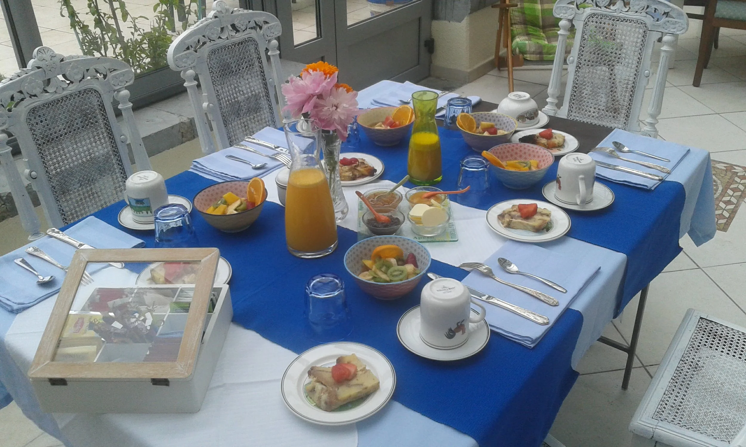 Table dressée pour un petit déjeuner avec des bols de fruits, des verres de jus d'orange, des tasses, des assiettes avec des tartines, des couverts, un vase avec des fleurs, et une nappe bleue.