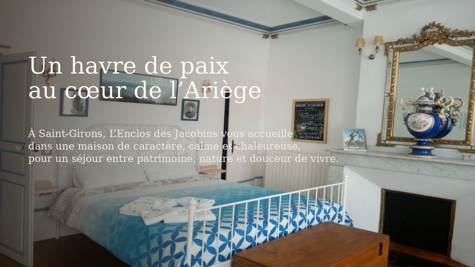 Chambre décorée dans un style classique avec un lit en fer blanc, un tapis à motif bleu, une cheminée avec une grande urne bleue et dorée, un miroir doré, des tableaux et une commode en bois.