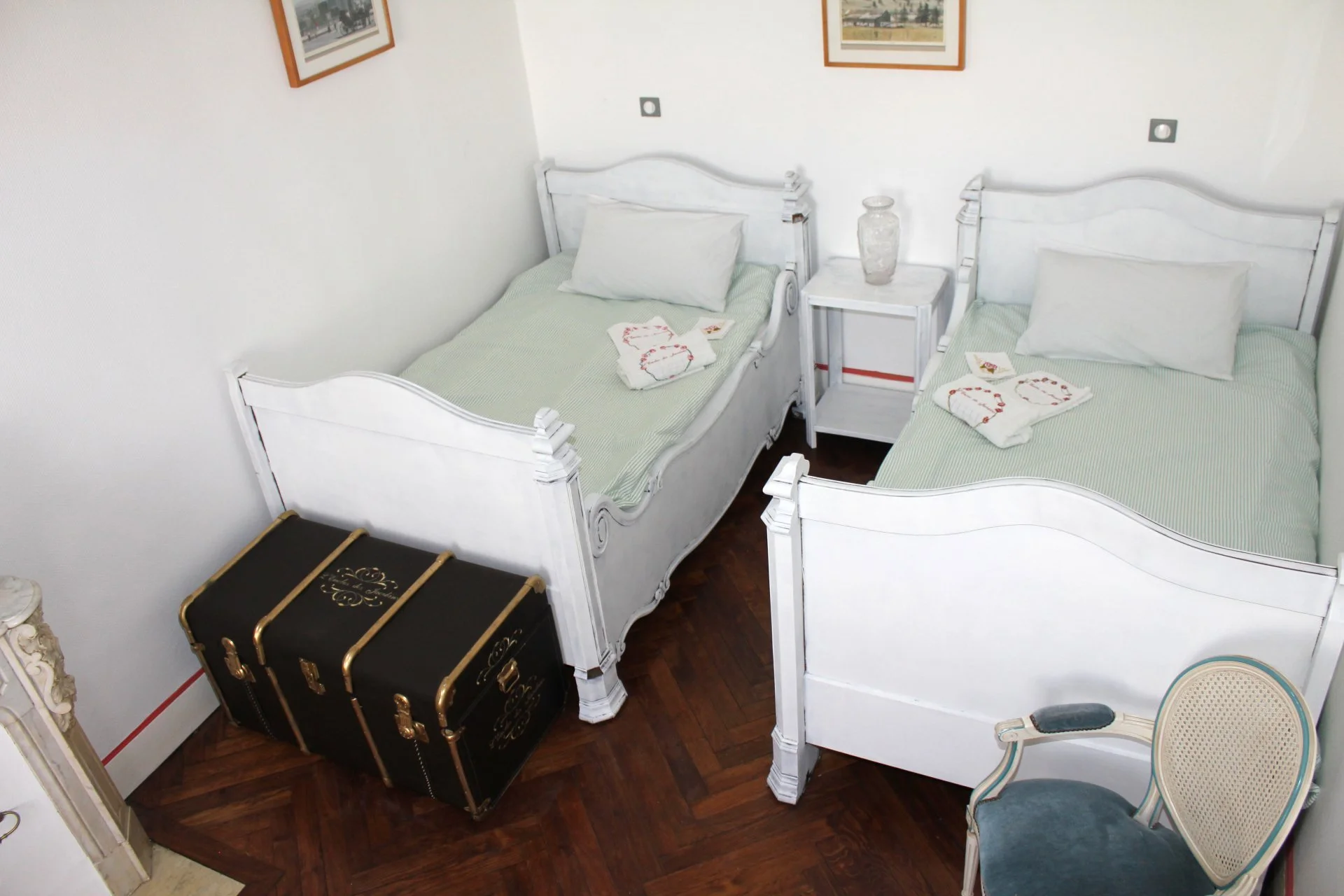 Chambre avec deux lits jumeaux en bois blanc, garlandés de draps verts et de coussins blancs, avec une petite table de nuit en blanc entre eux, une décoration de verres transparents, une malle noire et or au sol, deux tableaux encadrés au mur et deux chaises anciennes en tissu vert et ancien style.