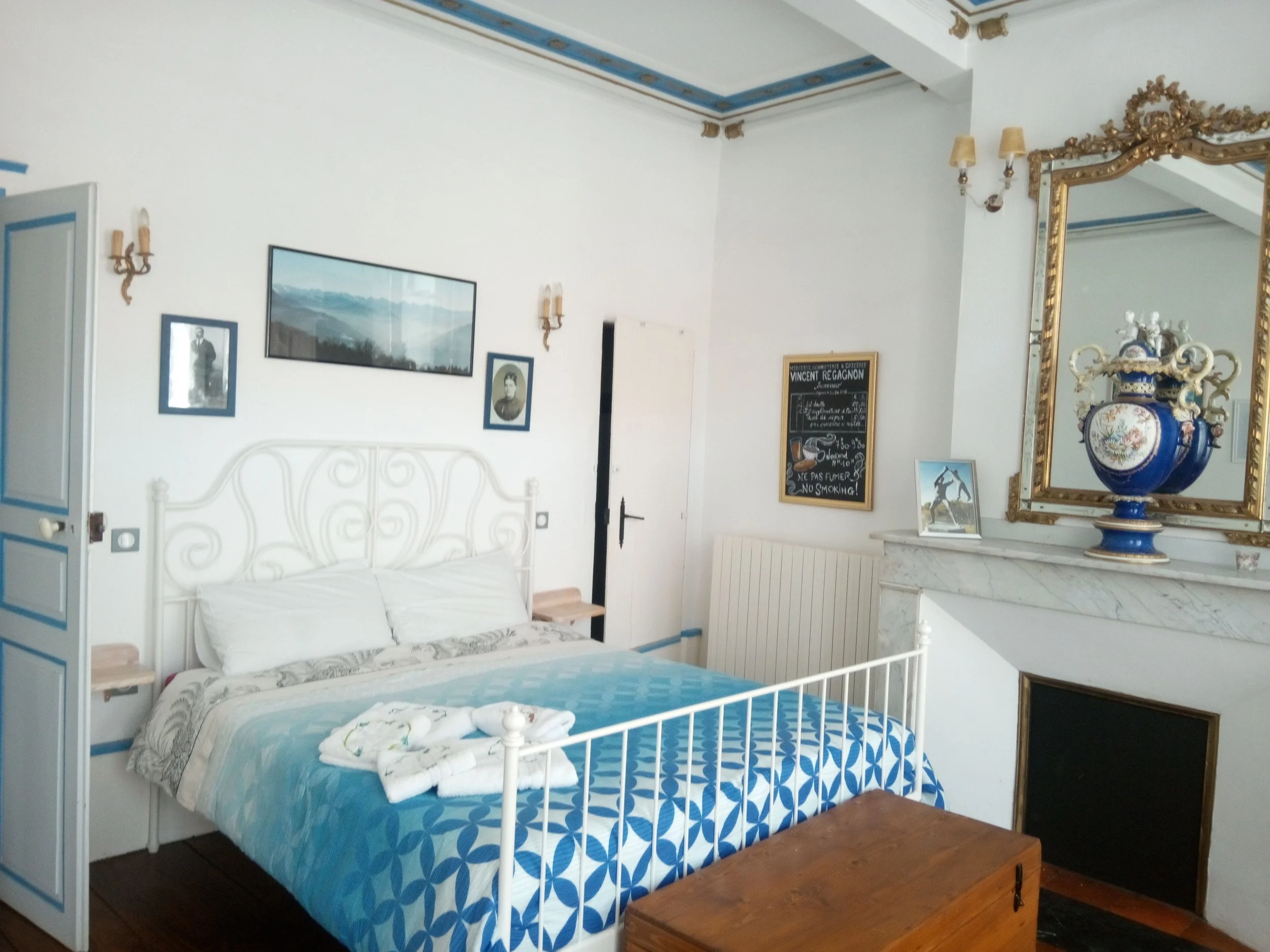 Une chambre avec un lit en fer blanc, couette bleue, et un mur décoré d'écrans et de cadres, un grand miroir doré, et une grande pièce de céramique avec des motifs floraux.