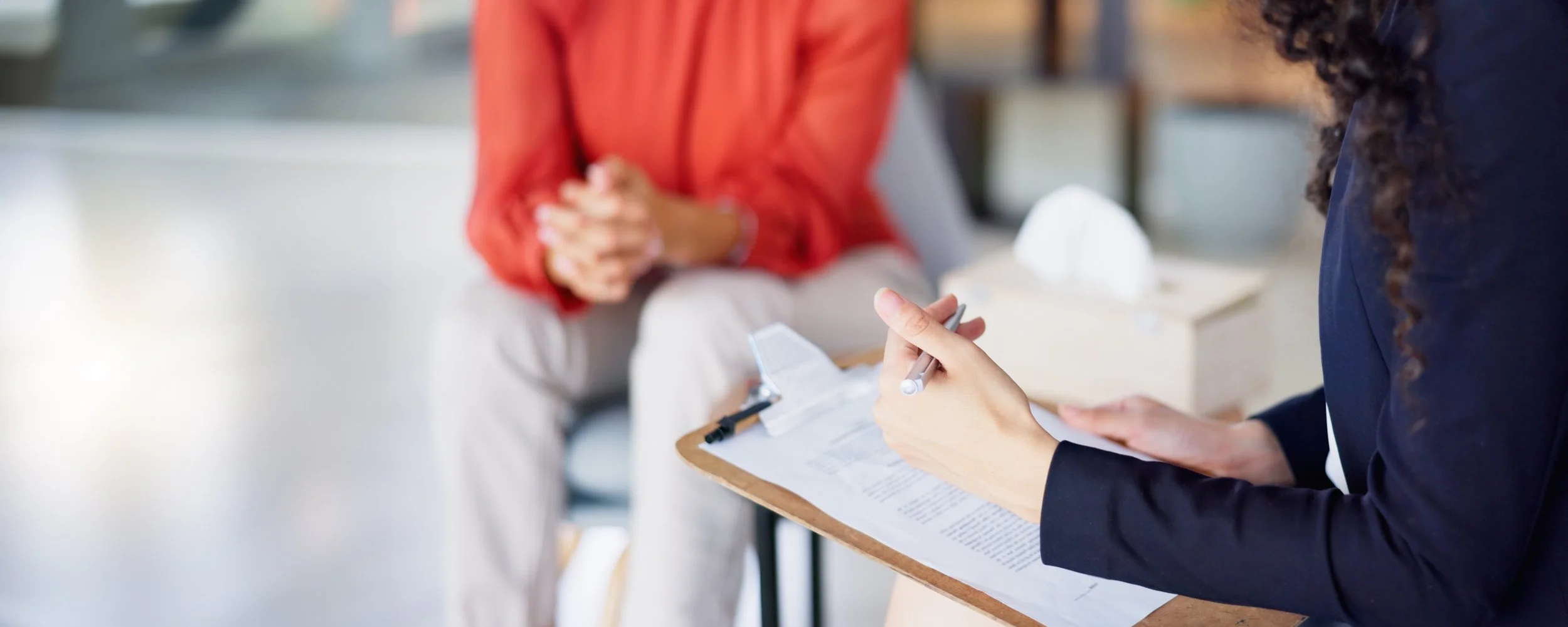 A person filling out a form on a clipboard while another person sits across, with both hands clasped. The setting appears to be an indoor office or waiting area, with a tissue box on a table in the background.