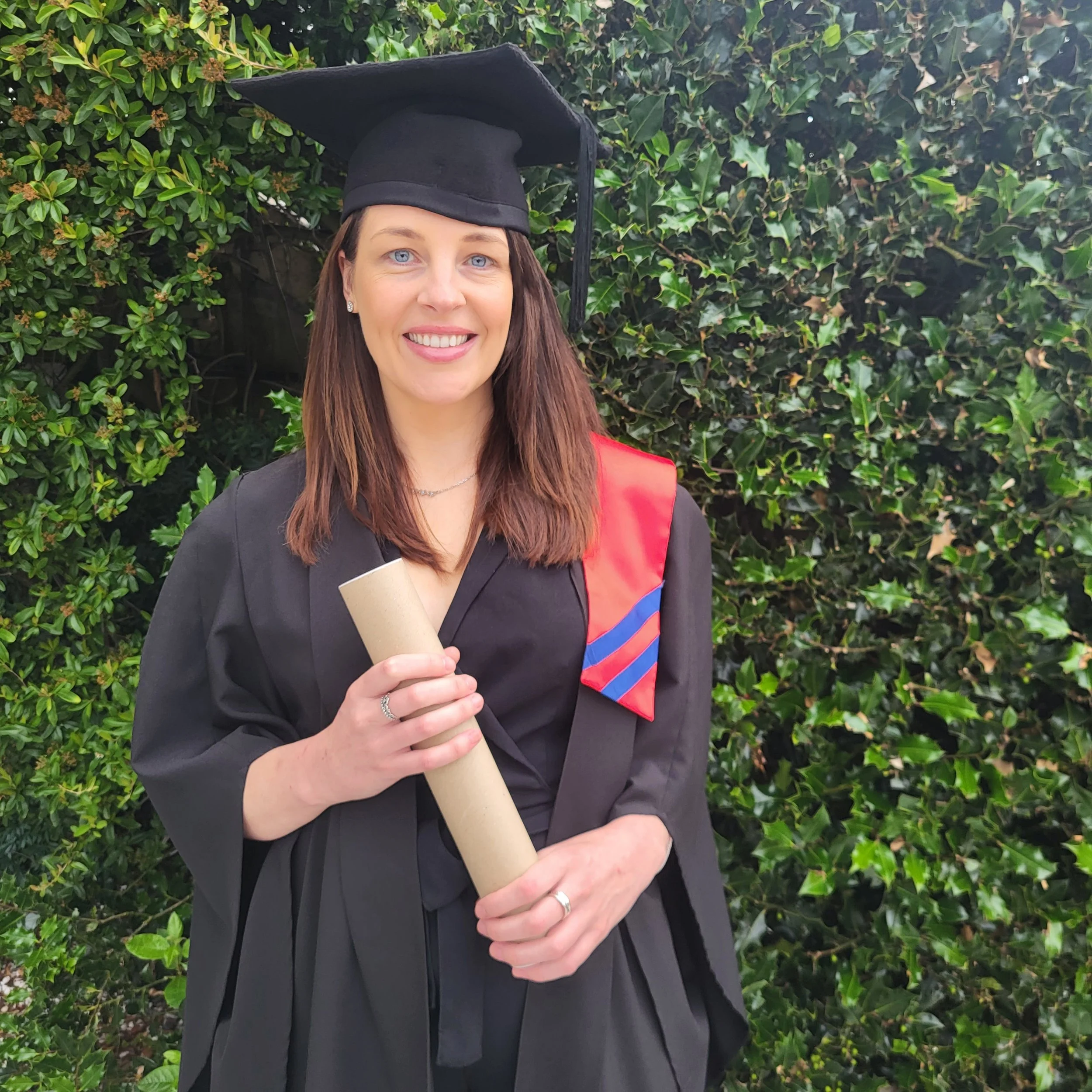 A woman in a graduation gown and cap smiling at the camera, holding a diploma, with green bushes in the background.