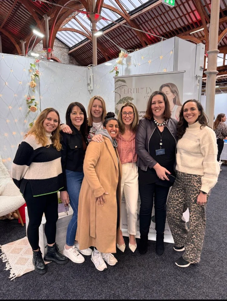 Group of seven women smiling and posing for a photo at an indoor event with decorative fairy lights and a banner that reads 'The Fertility'.
