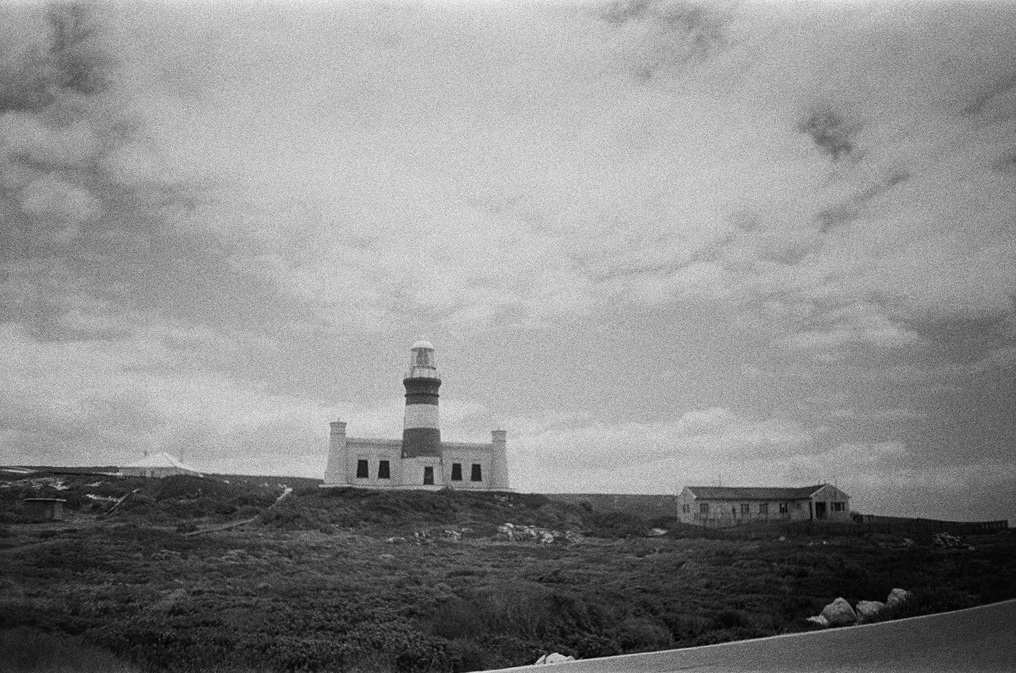 A black and white photo of a lighthouse on a small hill with a building at the base, and a sky filled with clouds in the background.