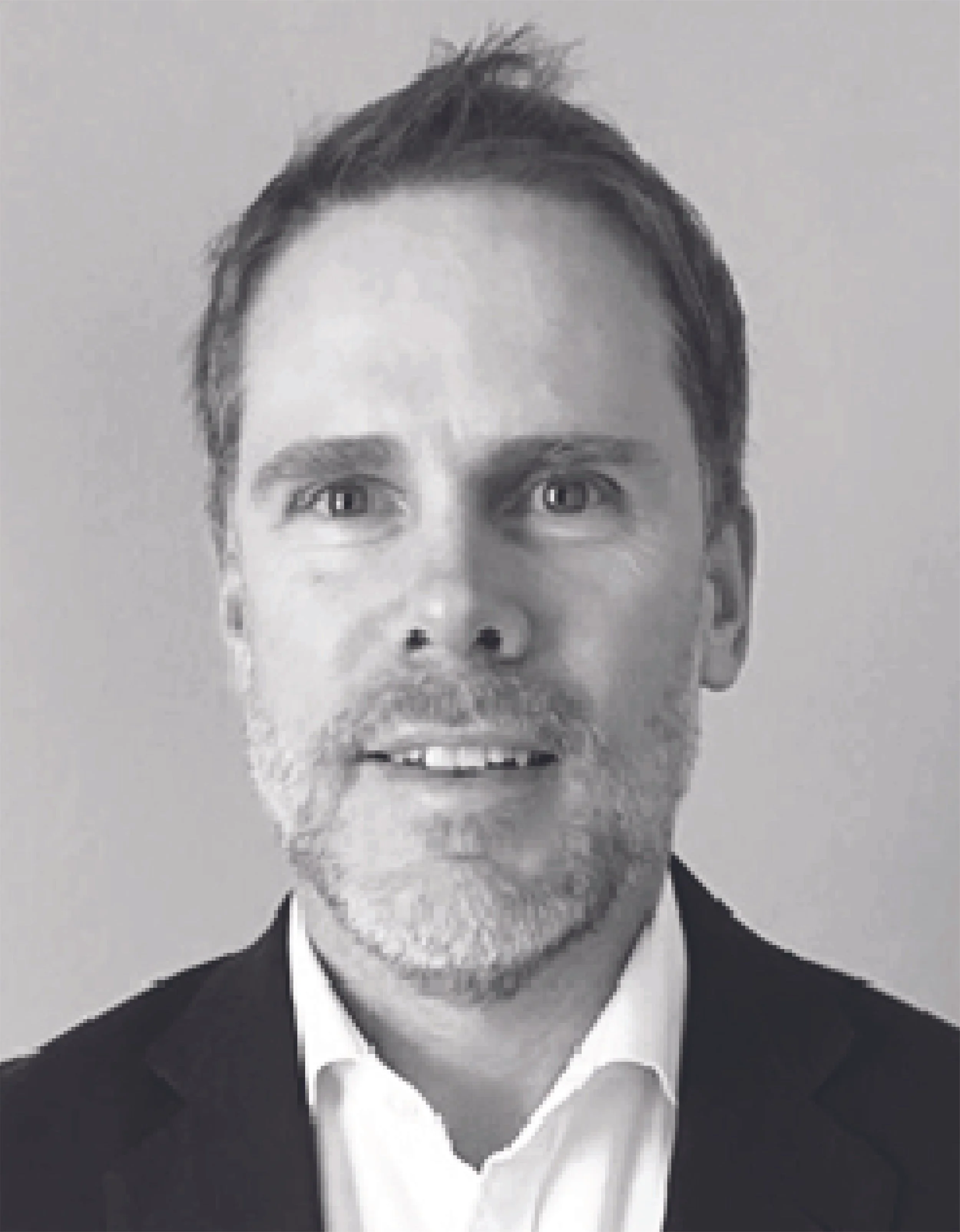 A black and white photograph of a man with a beard and mustache wearing a suit, shirt, and tie, with slicked-back hair, against a plain background.