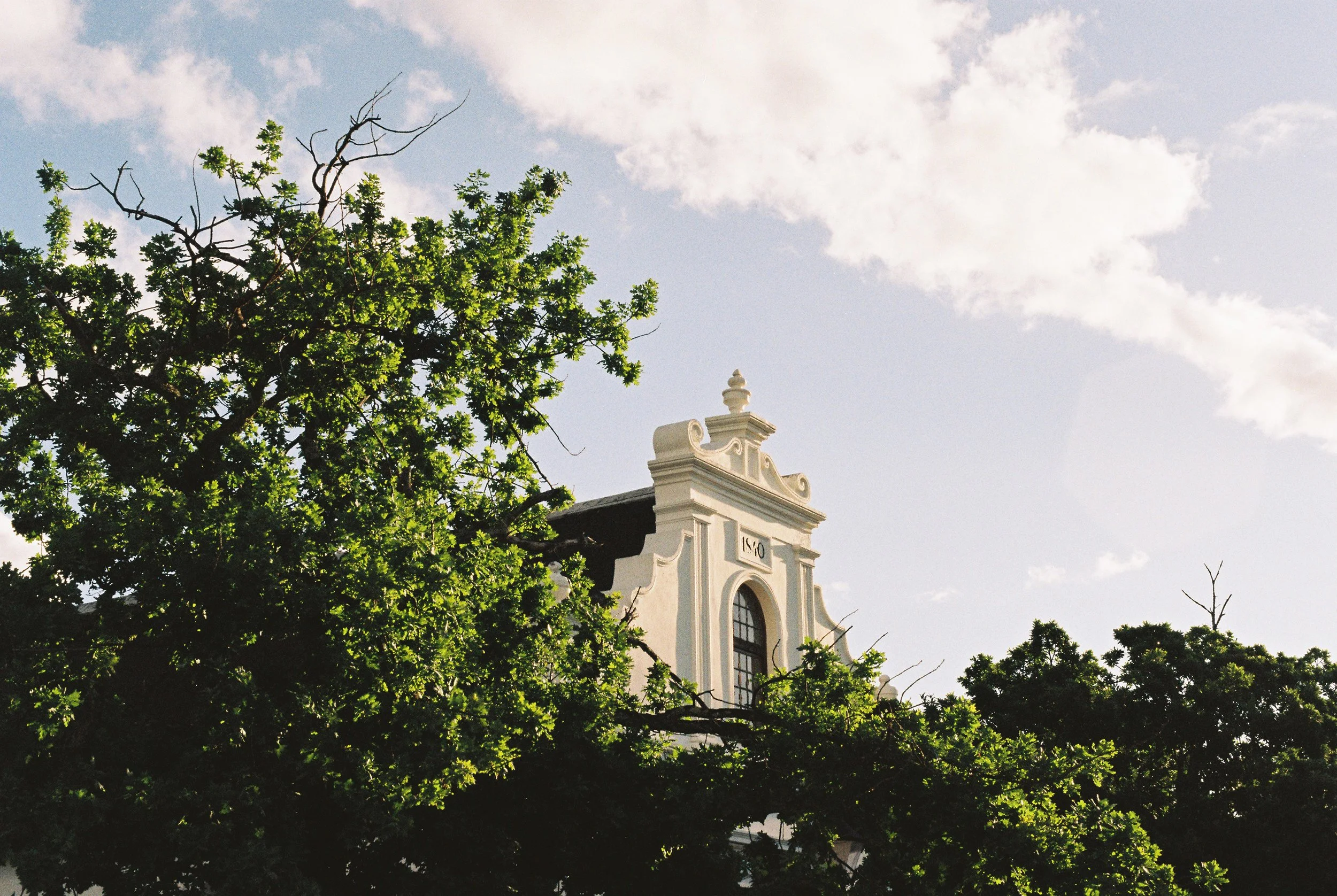 A white building with architectural details seen partly behind green leafy trees against a partly cloudy sky.