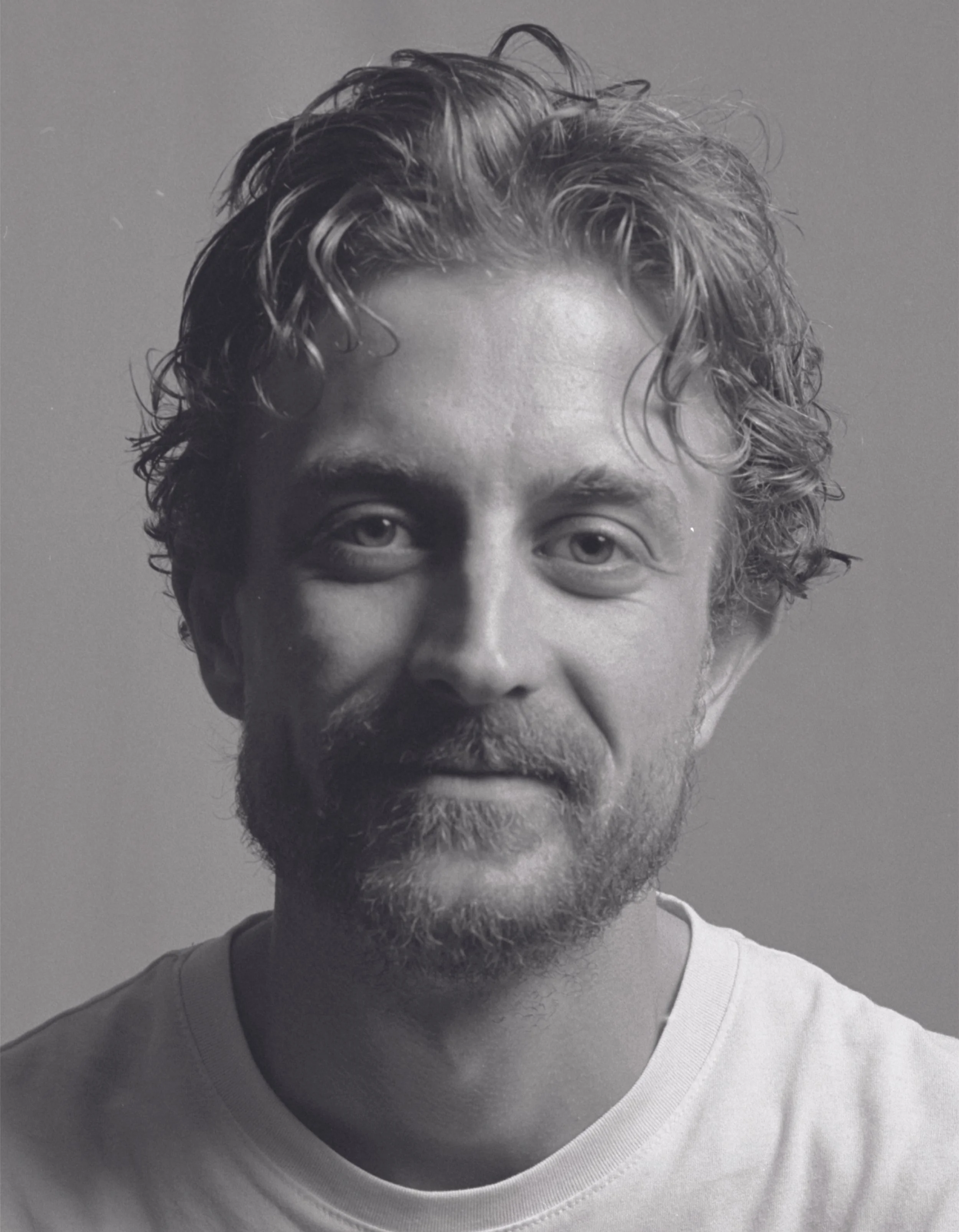 A black and white close-up portrait of a man with curly hair and a beard, wearing a white T-shirt.