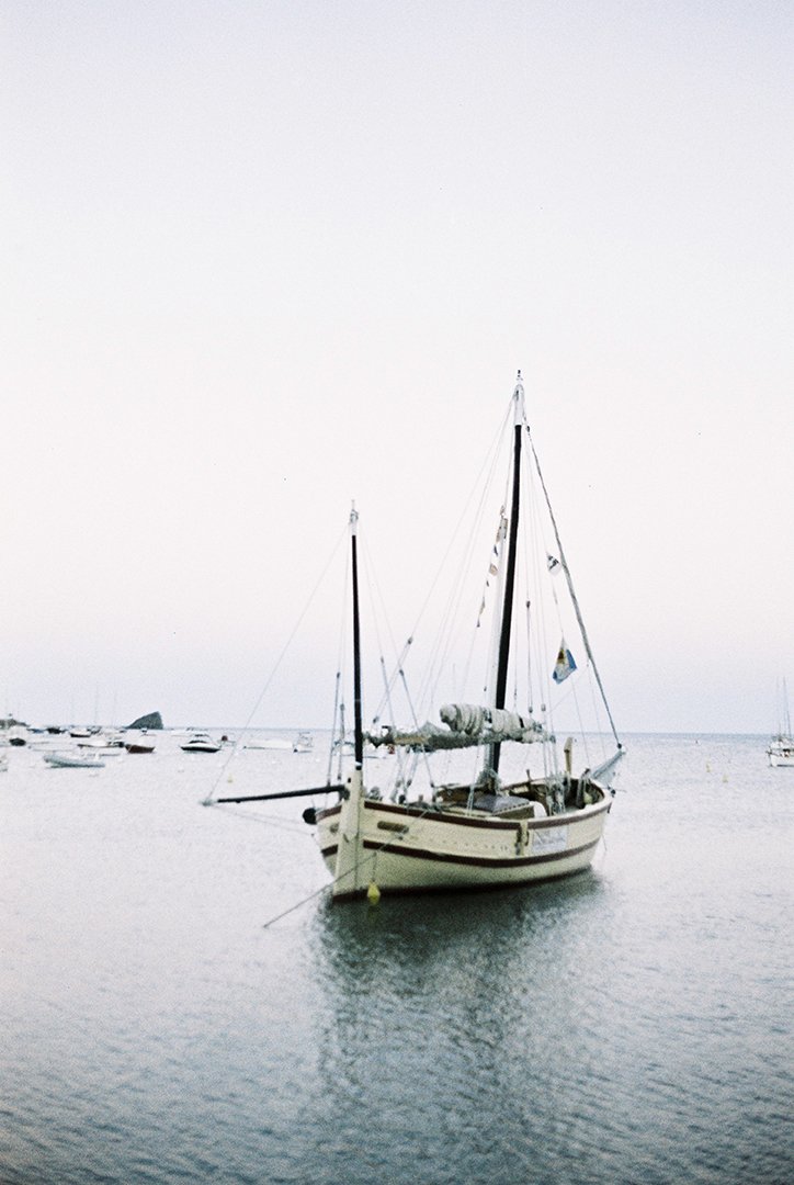 A sailboat anchored near a calm harbor with several smaller boats in the background, overcast sky.