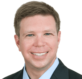A professional headshot of a man with short brown hair, smiling, wearing a dark blazer and light blue shirt against a white background.