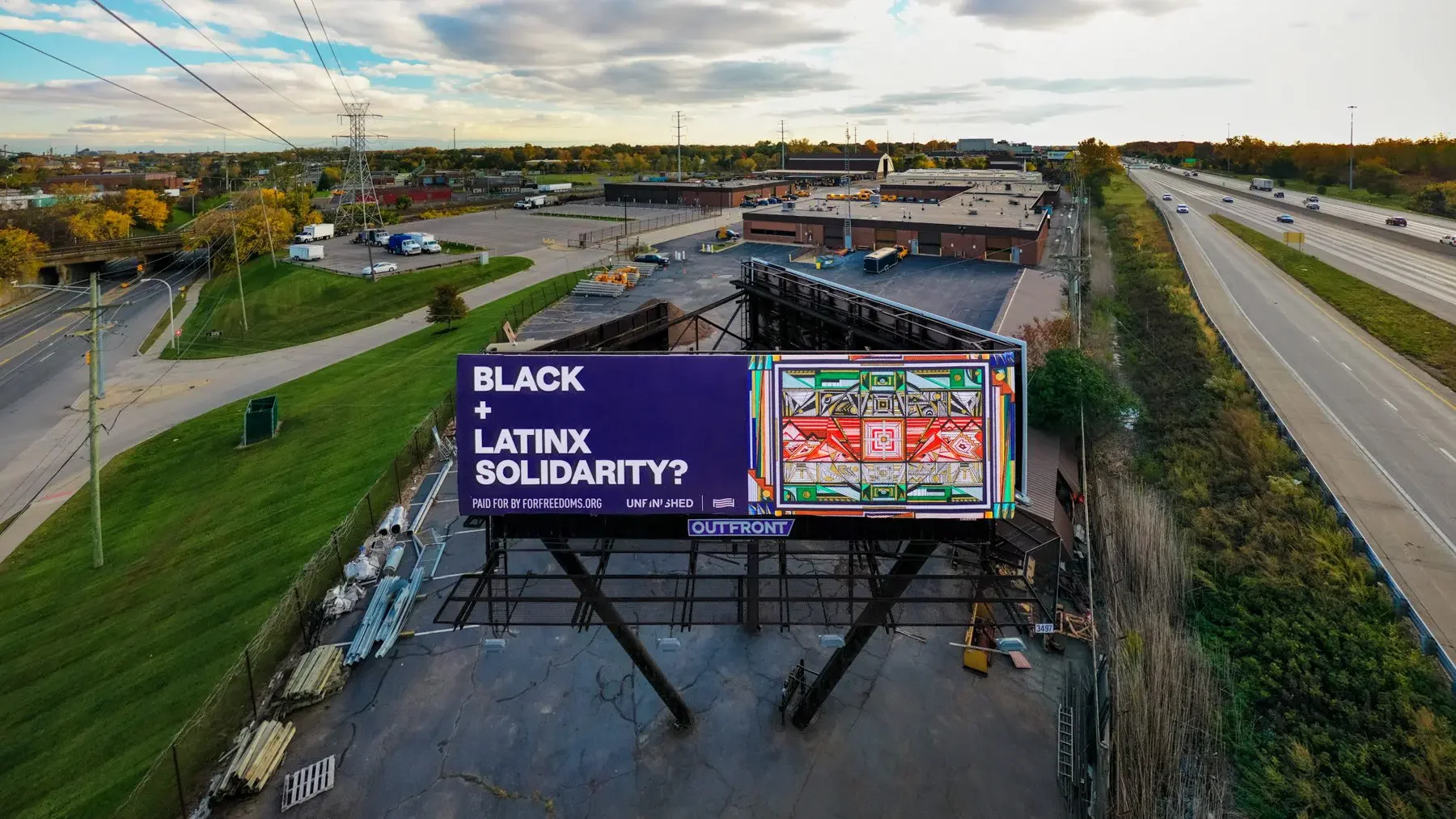 View from above, looking down on the billboard with the text "Black + Latinx Solidarity?" alongside Zahyr Lauren's artwork 'Black and Mexican Solidarity Flag'. In the background a highway and other roads are visible. 