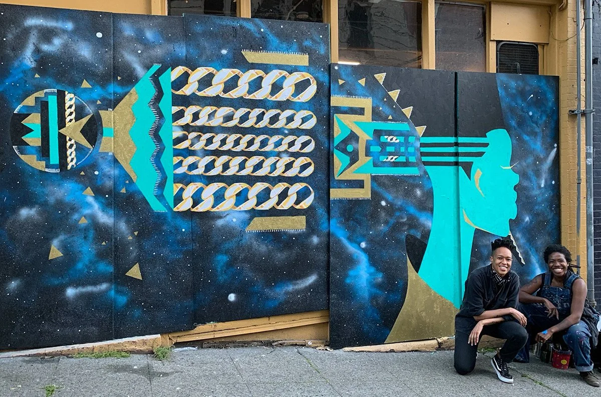 Photograph of two people posing in front of a large blue, turquoise and gold mural showing an afropick and a regal person in profile with a tall flat-top hairstyle.