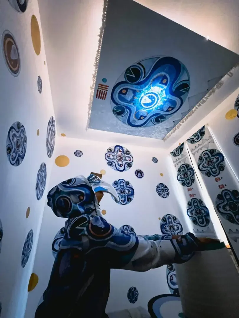 The artist Zahyr Lauren wears a futuristic hazmat suit and mask printed with their artwork 'Time Flow'. They are in a small room wallpapered with the artwork, looking up at a light in the ceiling, also decorated with the same art. 