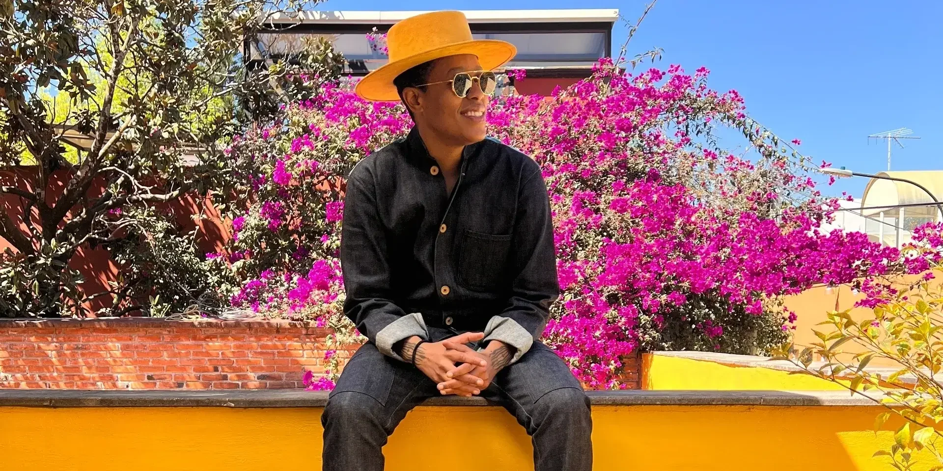 Photograph of Zahyr Lauren wearing black jeans and shirt with a yellow hat, sitting on a wall with pink flowers behind them.