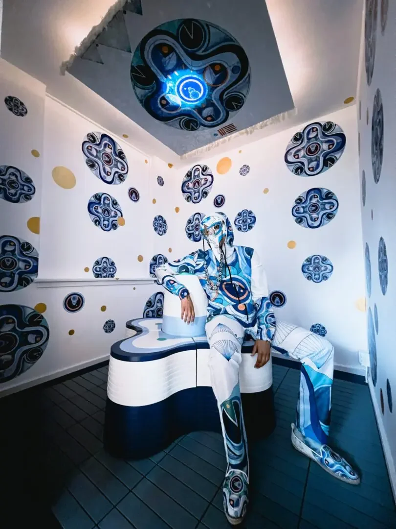 The artist Zahyr Lauren wears a futuristic hazmat suit and mask printed with their artwork 'Time Flow'. They sit in a small room wallpapered with the artwork. Above them, a light in the ceiling is also decorated with the same art. 