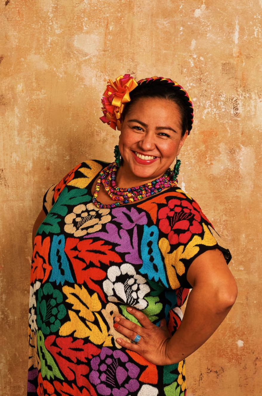 A smiling woman wearing colorful traditional Mexican attire, including a floral embroidered dress, vibrant jewelry, and a flower-adorned headband, standing against a textured beige background.