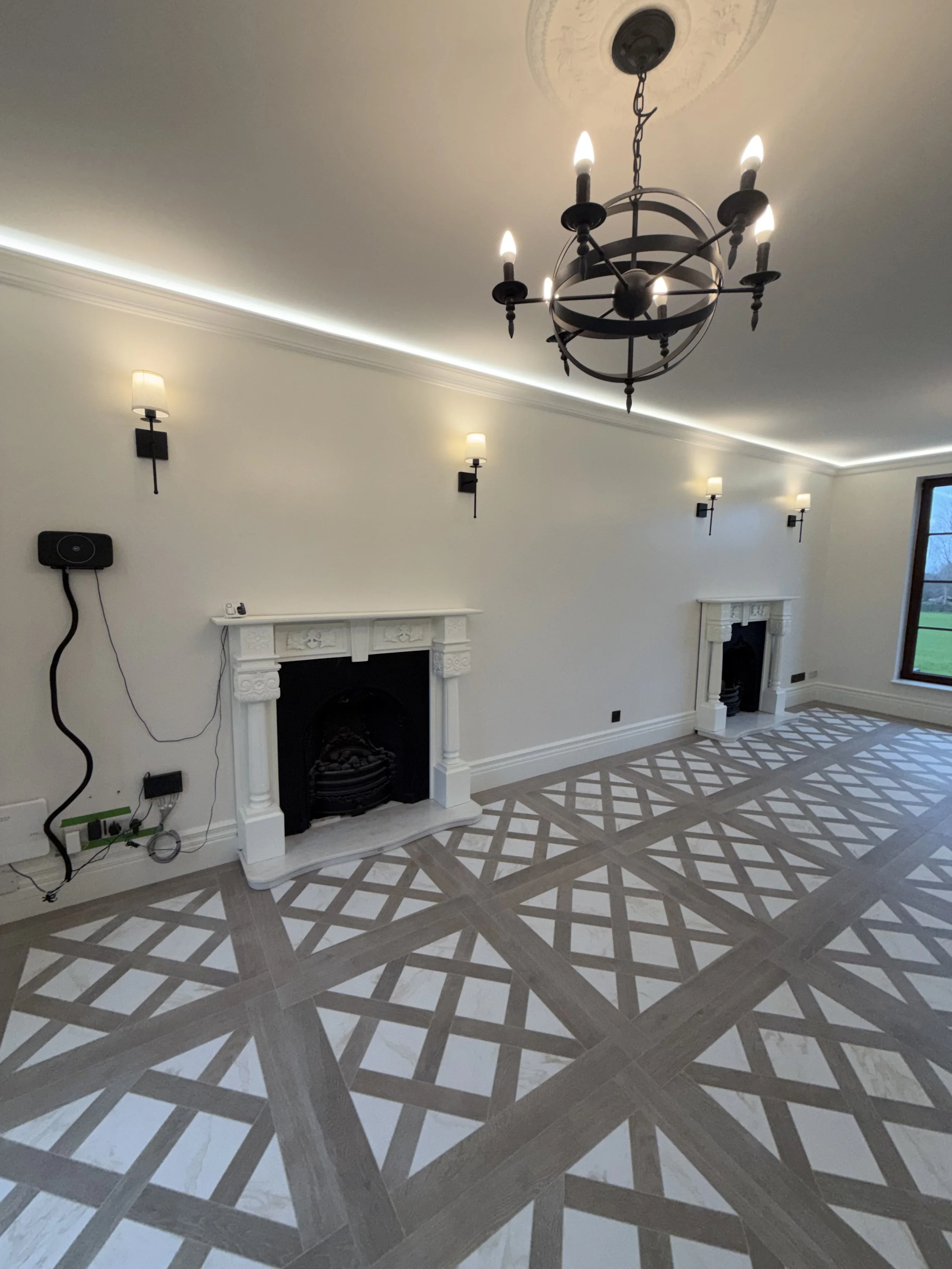 Empty living room with two white fireplaces, black accents, and a geometric patterned tile floor. Chandelier hangs from the ceiling, with multiple wall-mounted lights illuminating the room.