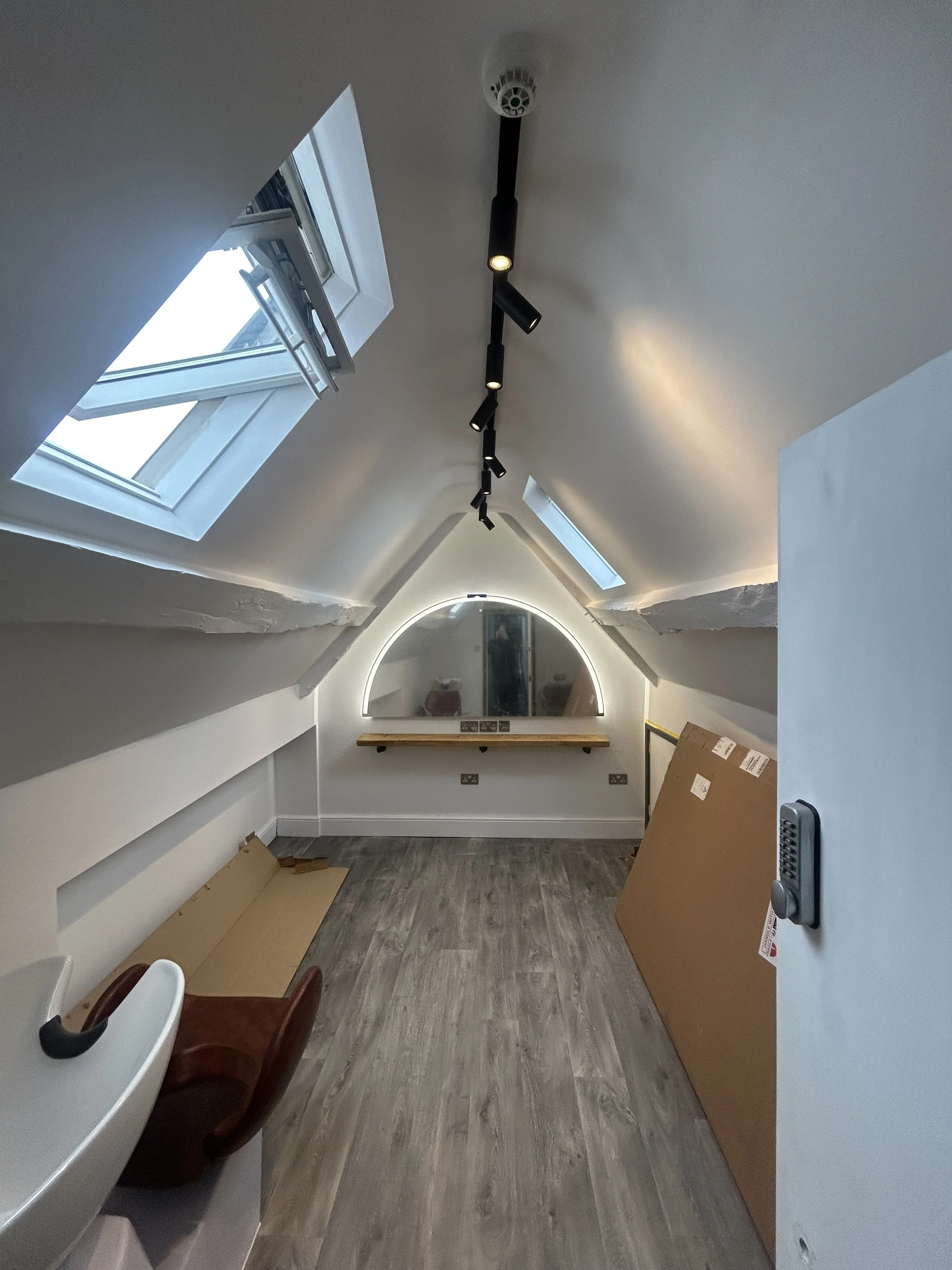 Attic room with two skylight windows, a large mirror with an arch-shaped LED light frame, a wooden shelf beneath the mirror, and a cardboard box leaning against the right wall with a small table and chair partially visible in the foreground.