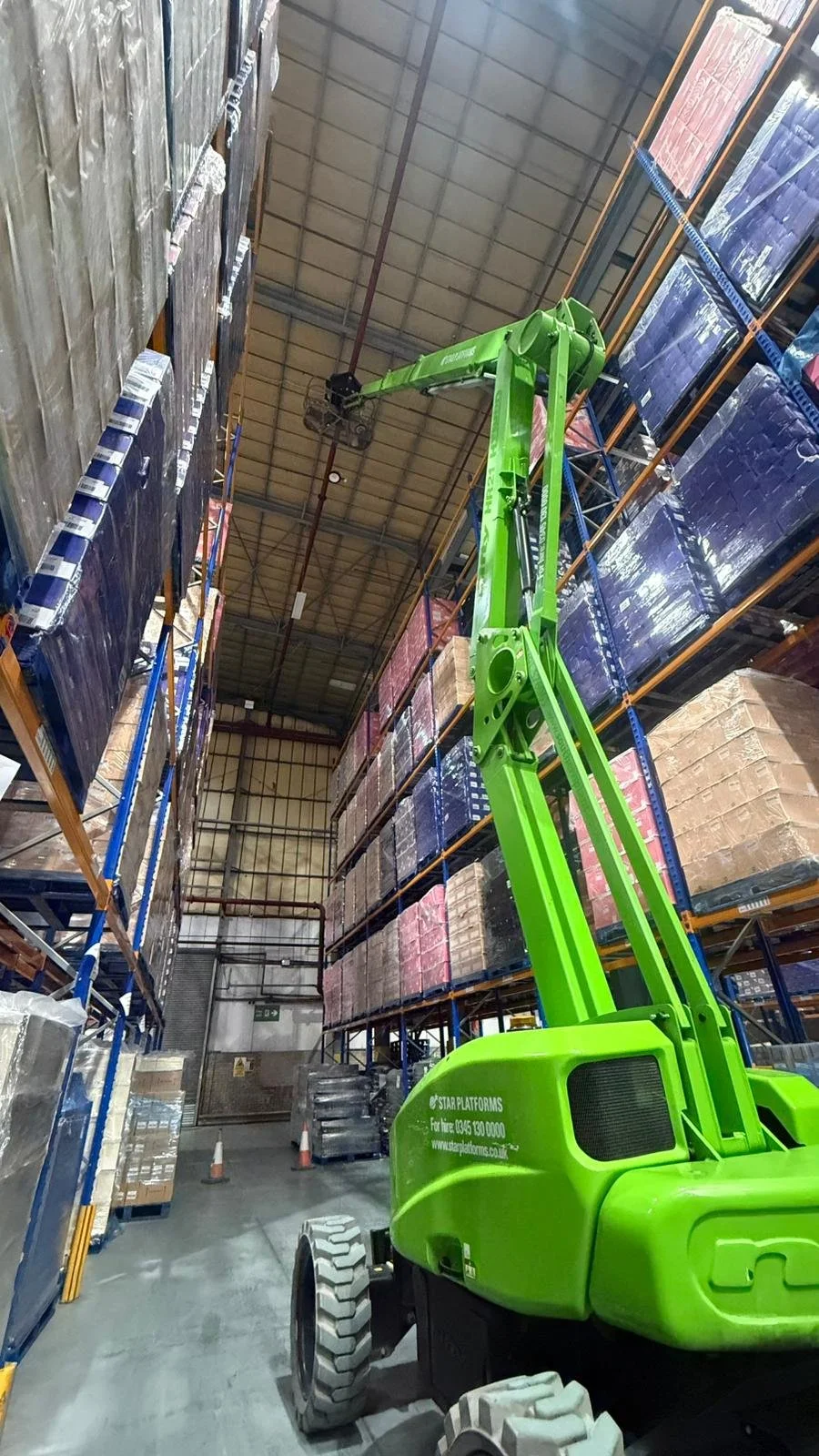 A green industrial reach forklift lifting items in a warehouse with tall shelves stacked with boxes.