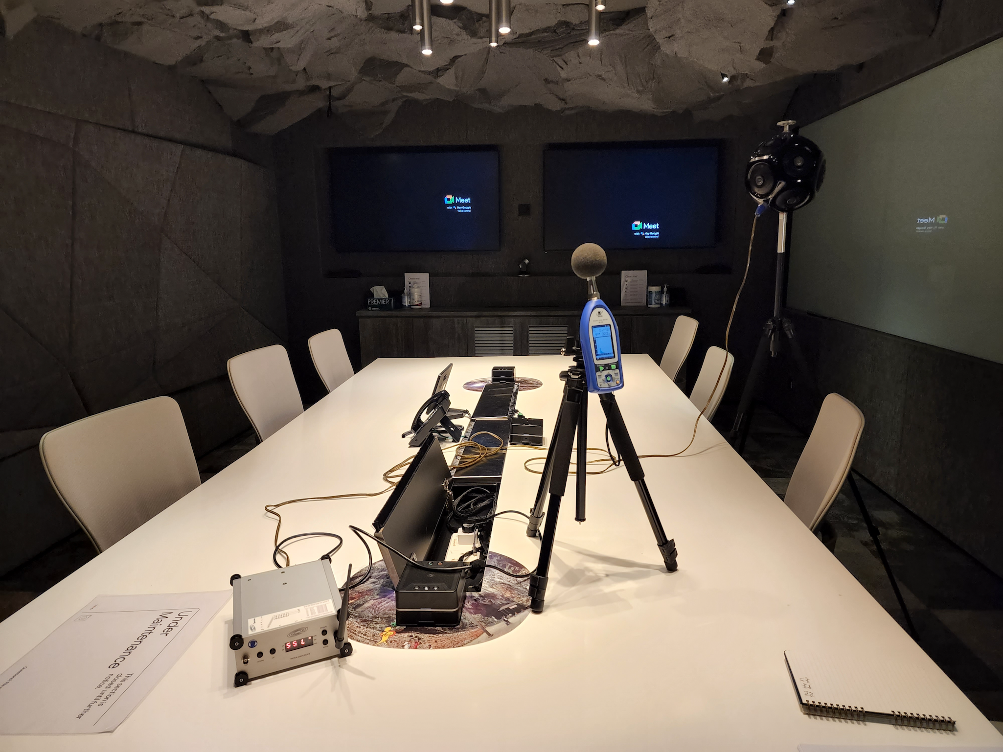 A conference room with a white table, multiple chairs, and various electronic equipment including a megaphone, a wireless microphone, and electronic control devices. There are two large screens on the wall displaying the Google Meet logo.