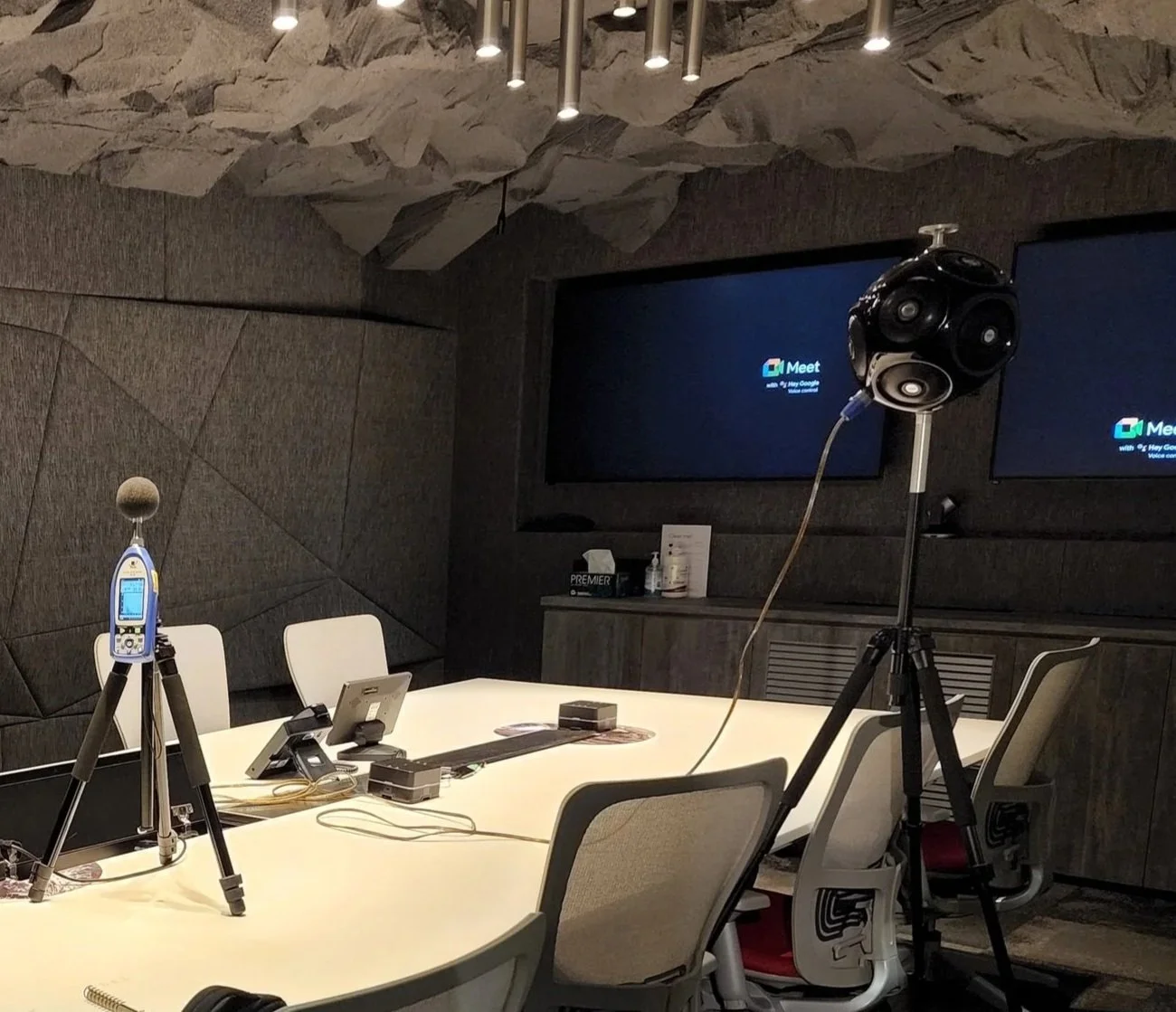 A room with a patterned black and white wall, a microphone on a stand, a computer monitor, and several posters on the wall. One person wearing a face mask and striped shirt is visible in the background.
