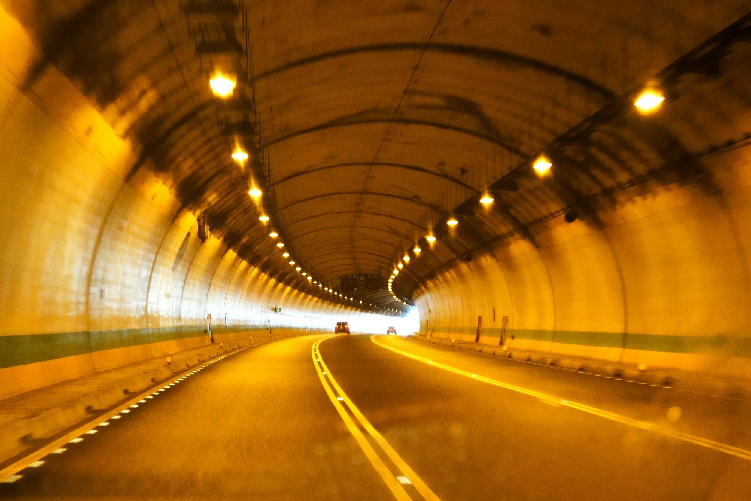 A view inside a tunnel with a curved ceiling, illuminated by yellowish lights. The road has a double yellow line in the center, with a few cars driving through toward the dark exit of the tunnel.