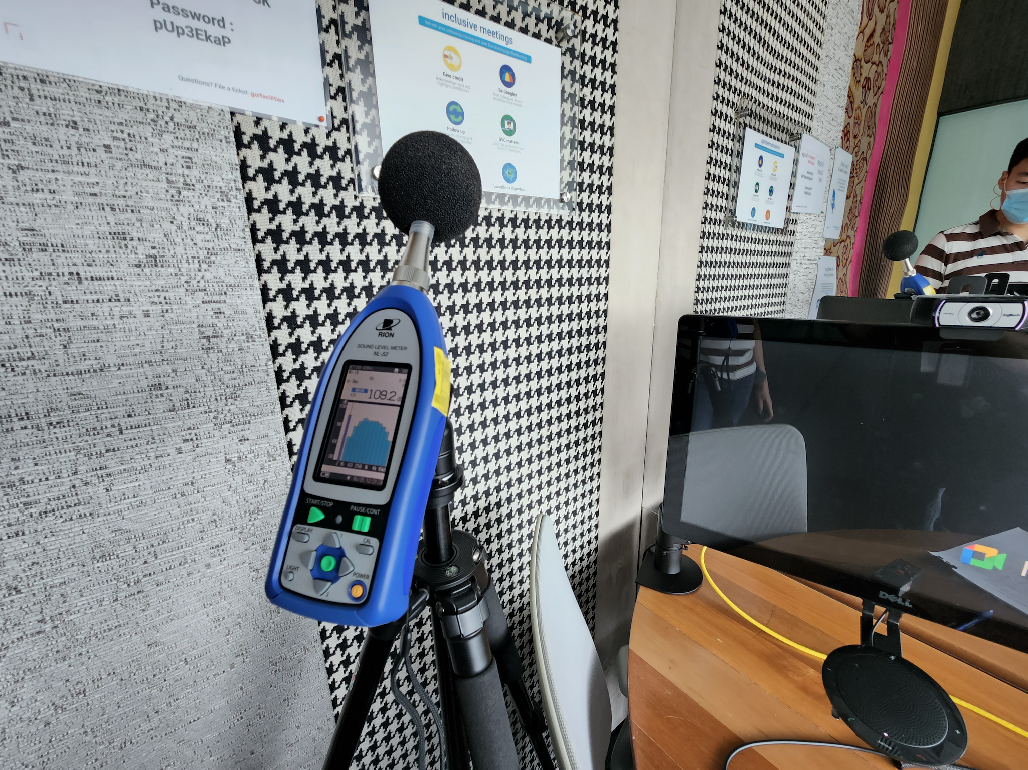 A room with a patterned black and white wall, a microphone on a stand, a computer monitor, and several posters on the wall. One person wearing a face mask and striped shirt is visible in the background.