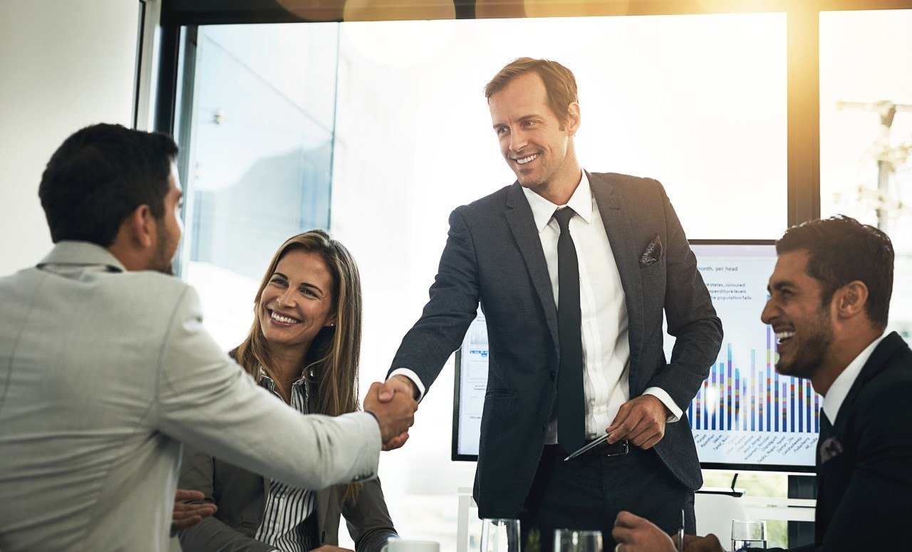 Business meeting with four professionals, two men and two women, shaking hands in a conference room with a presentation screen displaying graphs in the background.