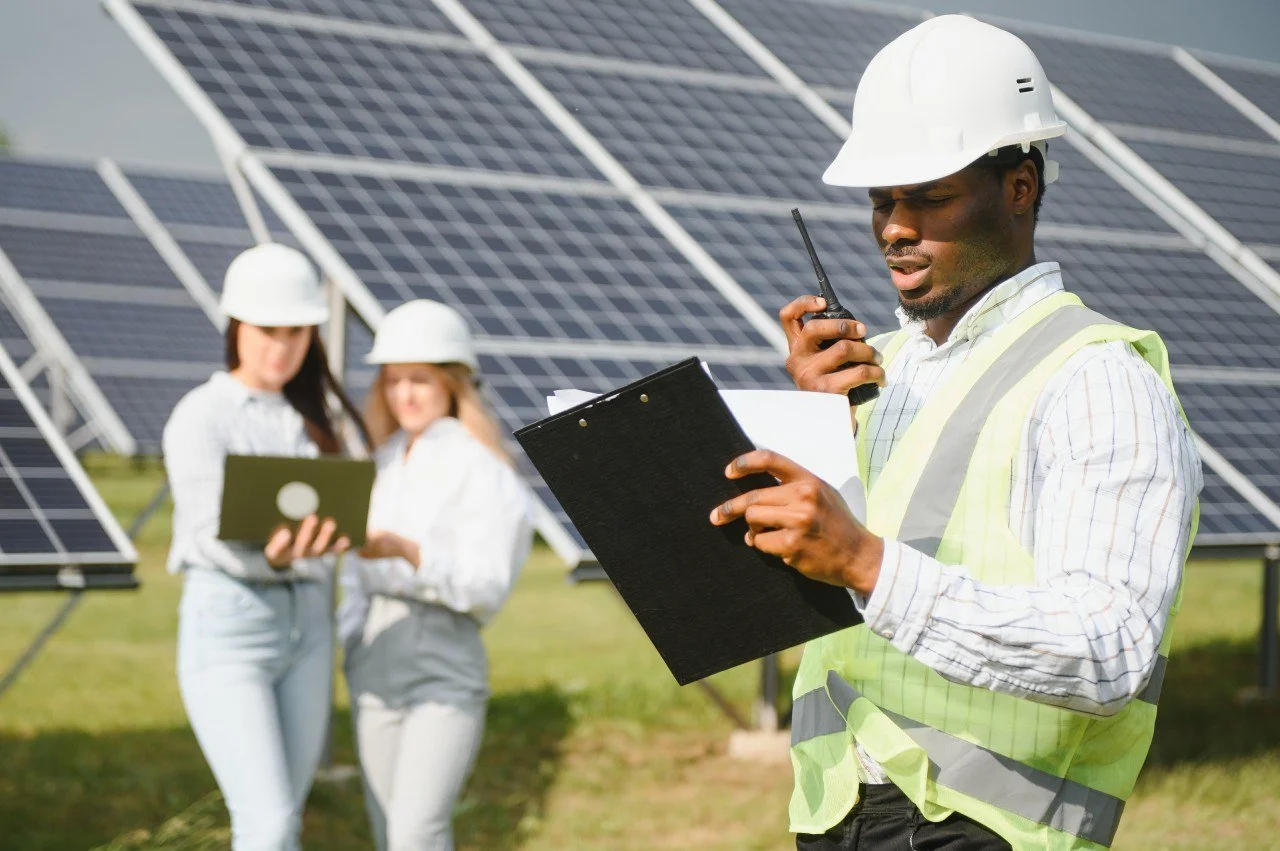 A man in a white safety helmet and reflective vest reviewing documents with a walkie-talkie at a solar panel installation site, with two women in safety helmets and casual attire in the background, also examining documents near solar panels.