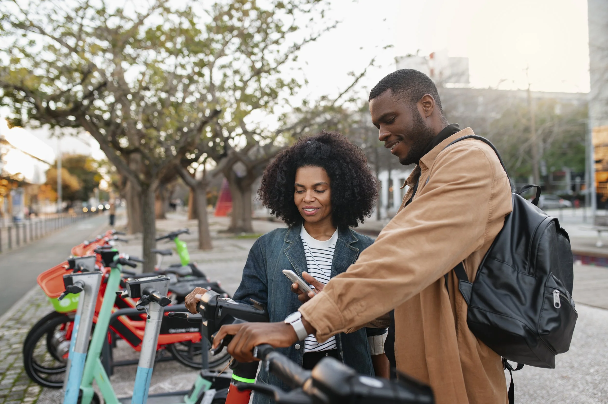 Two people, a man and a woman, are standing outdoors next to a row of electric scooters. They are looking at a smartphone together, smiling, with a background of trees, bicycles, and a sidewalk.