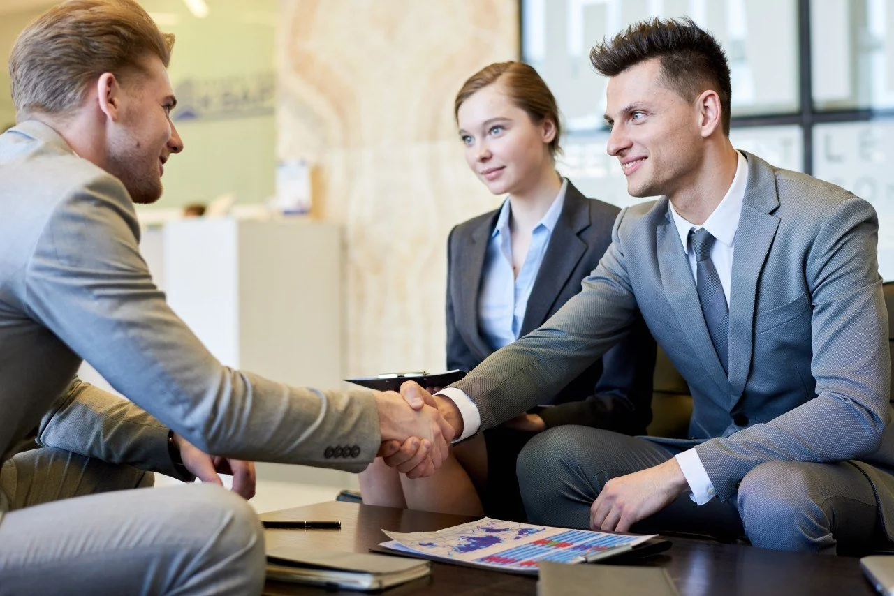 Two men in suits shaking hands across a table, with a woman in a suit sitting beside them, in a bright office setting.