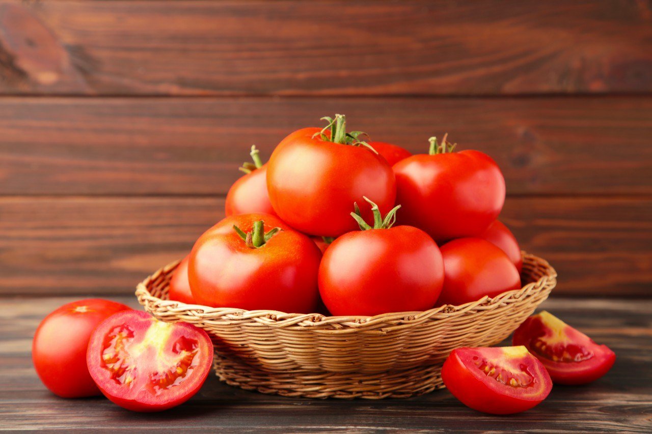 A basket filled with ripe red tomatoes on a wooden surface with a wooden background, some tomatoes sliced open showing the interior.