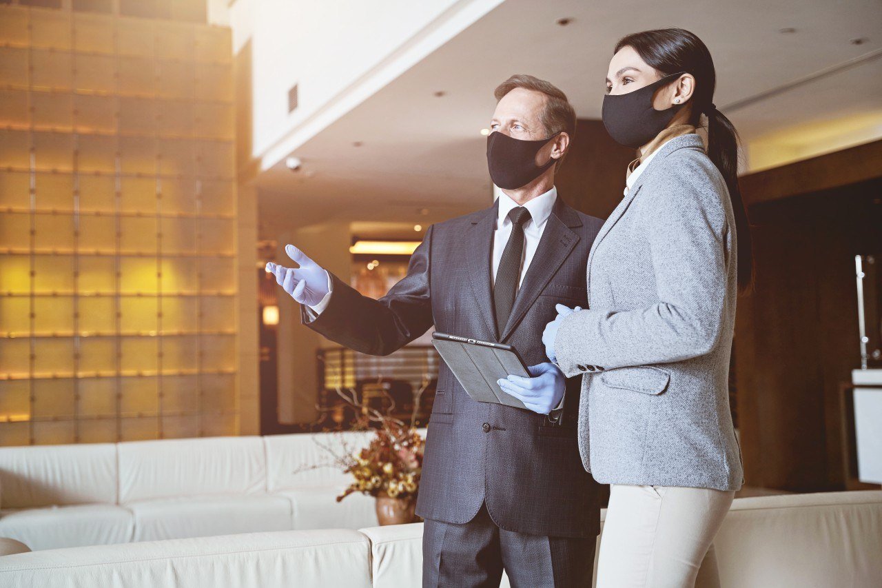 A man and woman wearing face masks and gloves, engaged in a discussion in a hotel lobby or reception area. The man holds a clipboard and is gesturing as he speaks.