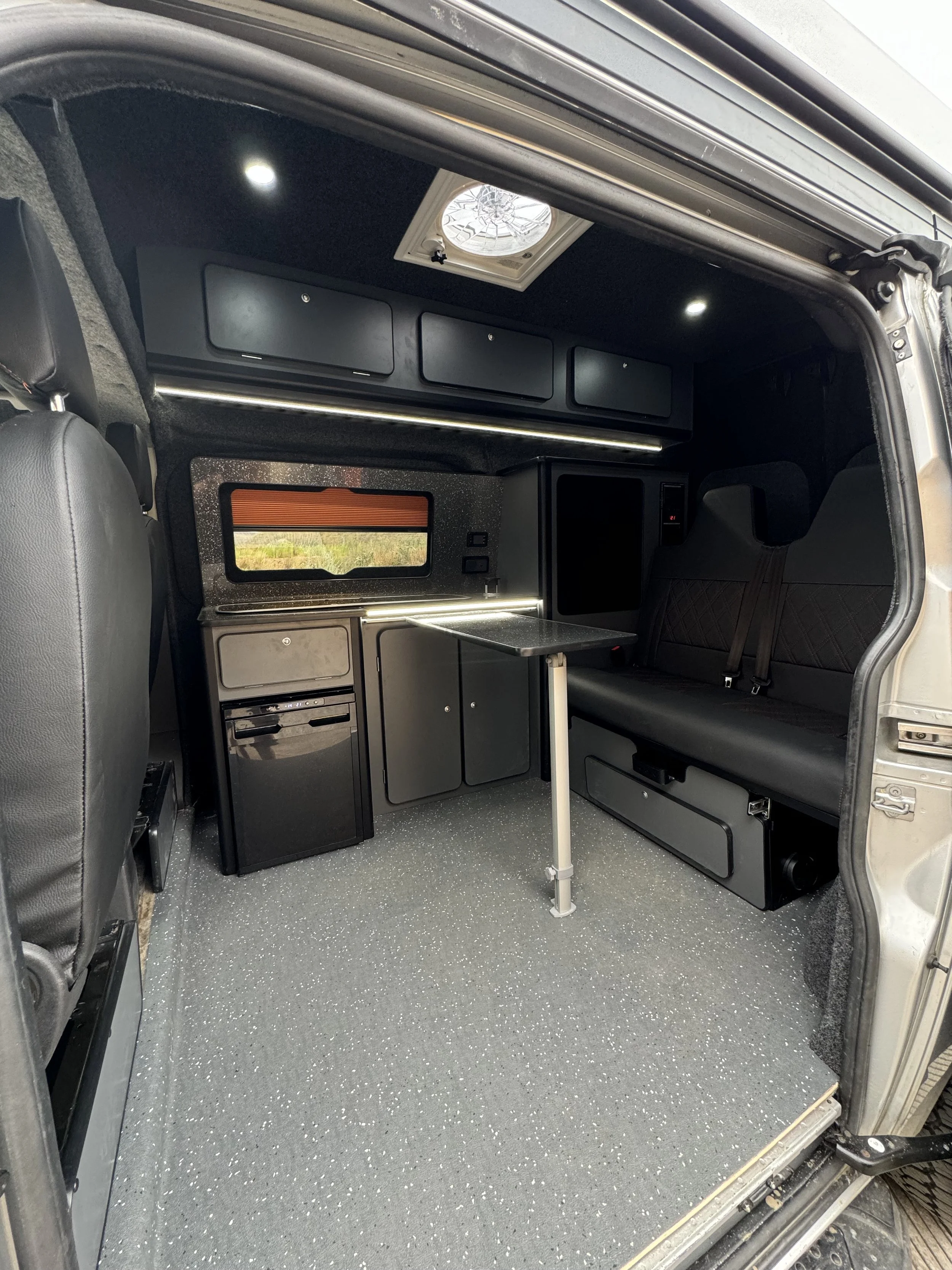 Interior of a campervan with black cabinets, a small table, a rock n roll bed with seat belts, a window with an orange blind, a small fridge, a ceiling vent, LED lights, carpet lined van.
