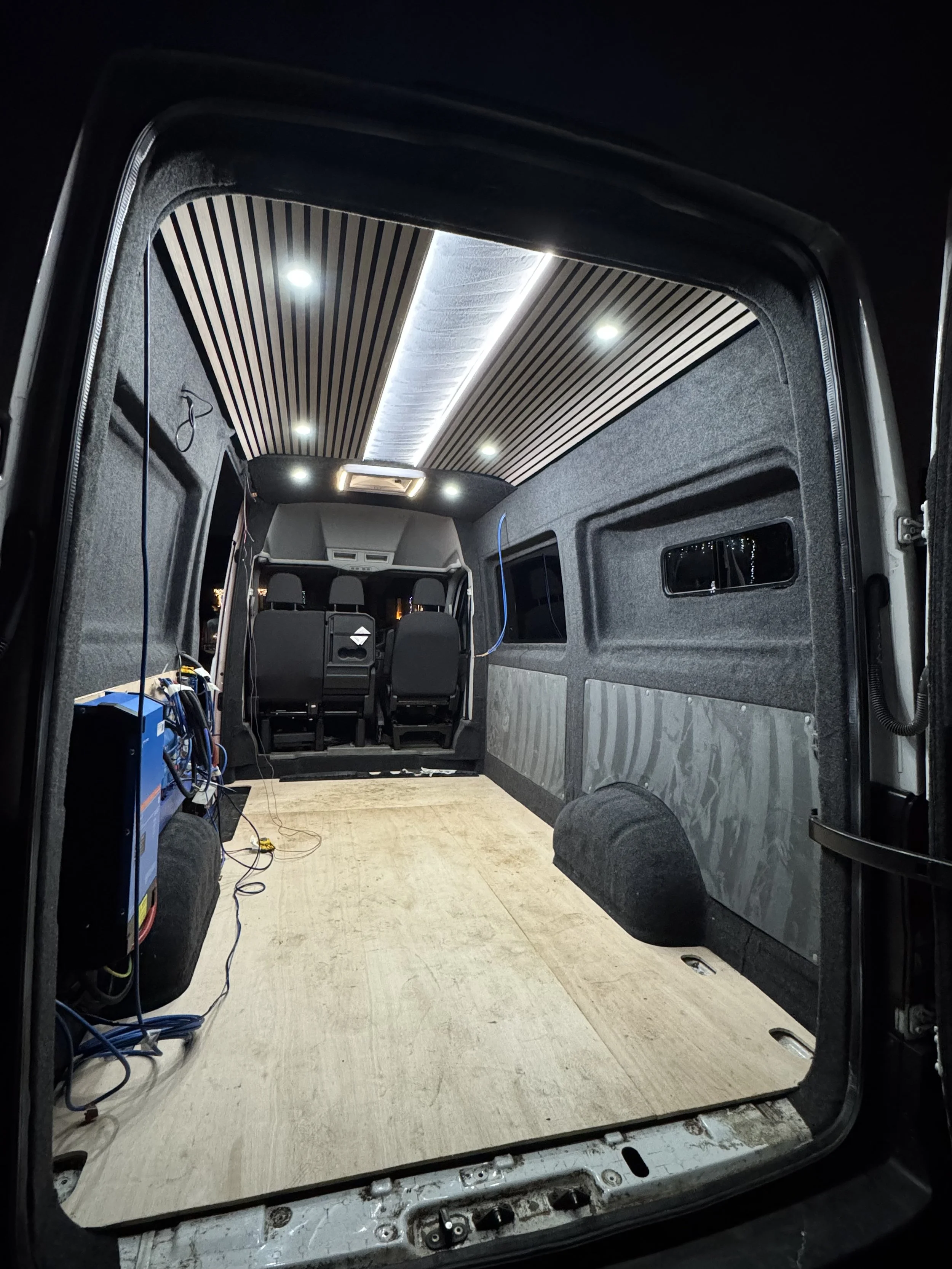 The interior of a van being renovated with new wood flooring, soundproof panels, and ceiling lights installed.