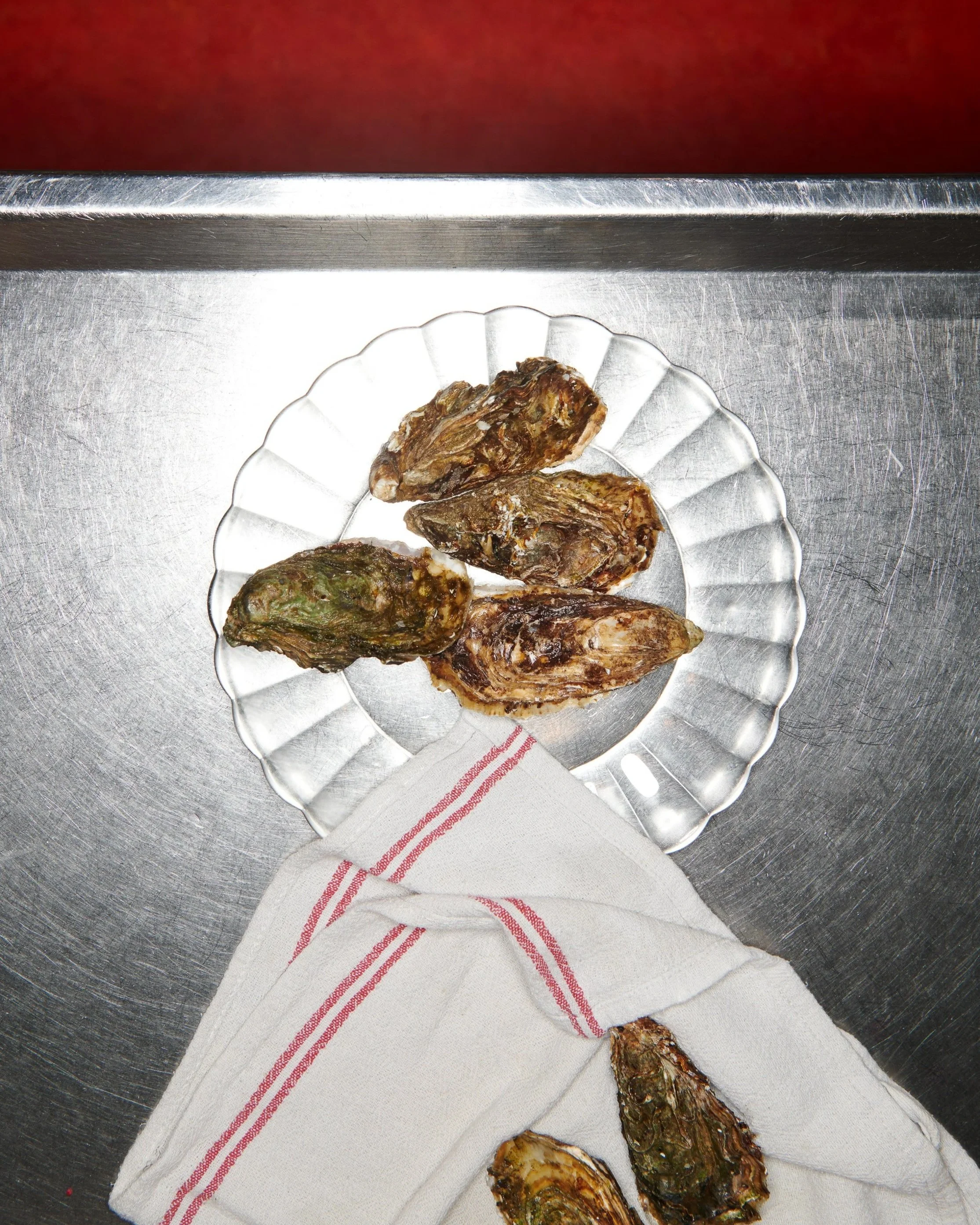 Gros plan sur une assiette en verre contenant des huîtres, posée sur une serviette blanche avec des rayures rouges, sur une surface métallique.