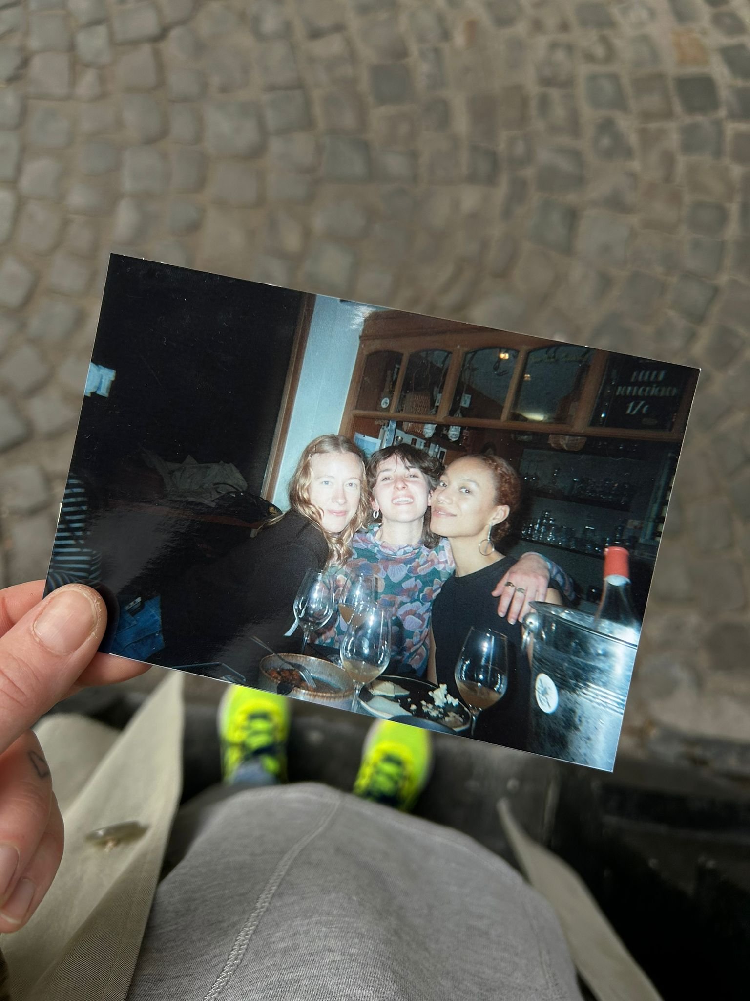 Une personne tient une photo d'un groupe de trois femmes souriantes assises à une table dans un restaurant ou bar, avec des verres et une bouteille de vin visible.