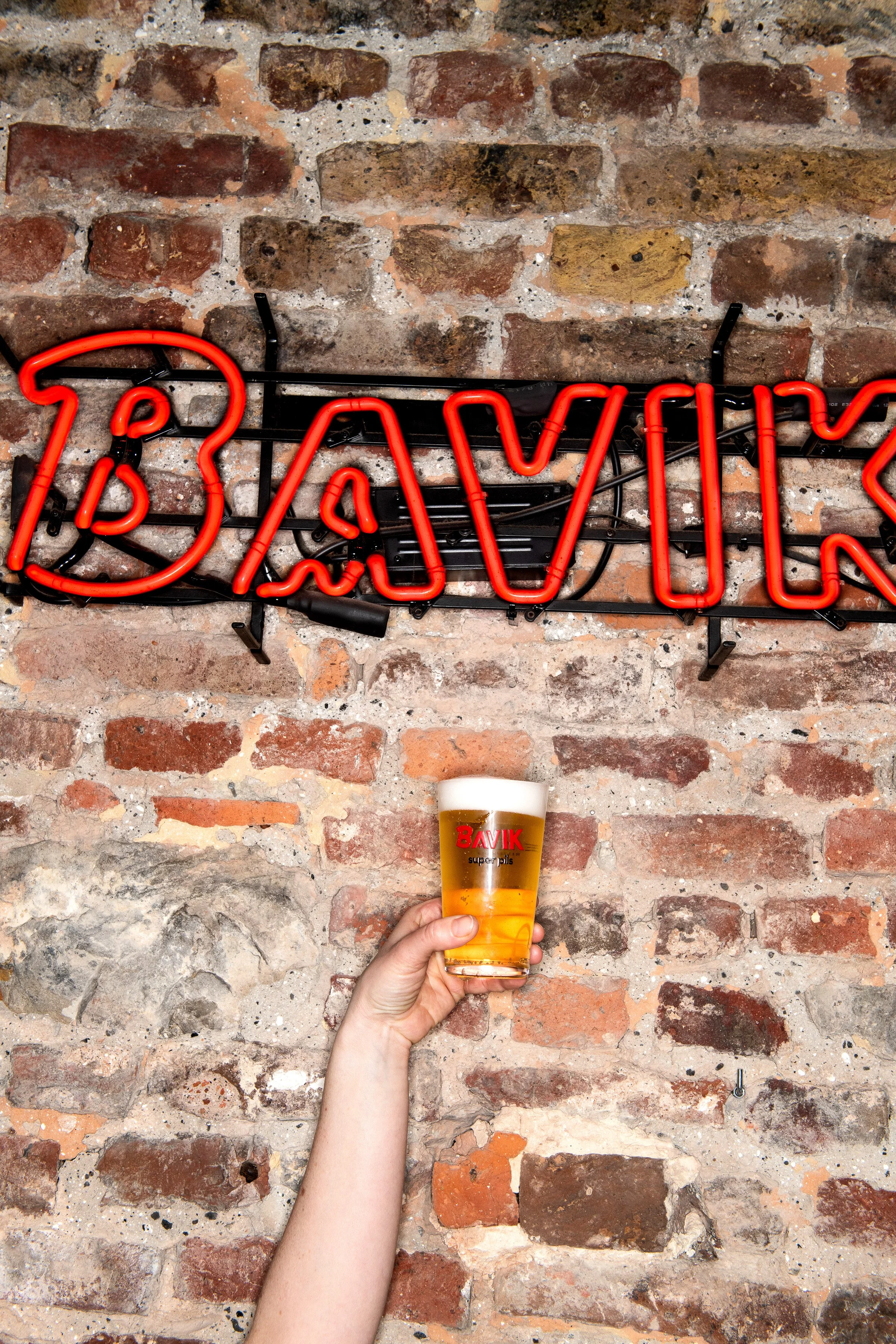 Une main tient un verre de bière blonde devant un mur en briques avec une enseigne lumineuse rouge et noire indiquant 'BAR'.